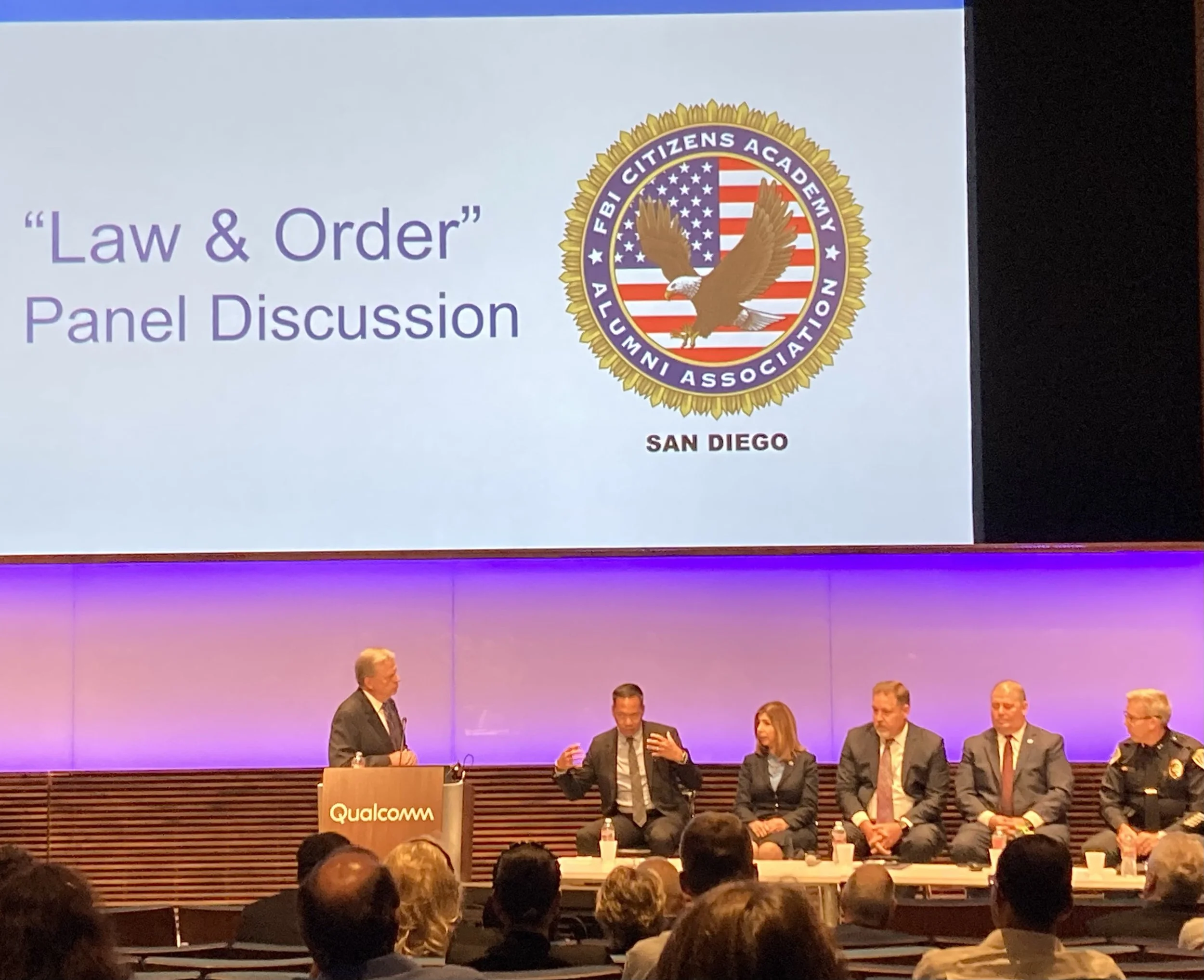 Panel discussion at a conference with a large screen displaying 'Law & Order' Panel Discussion and the FBI Citizens Academy Alumni Association logo, with six individuals seated on stage and a speaker at a podium labeled Qualcomm.