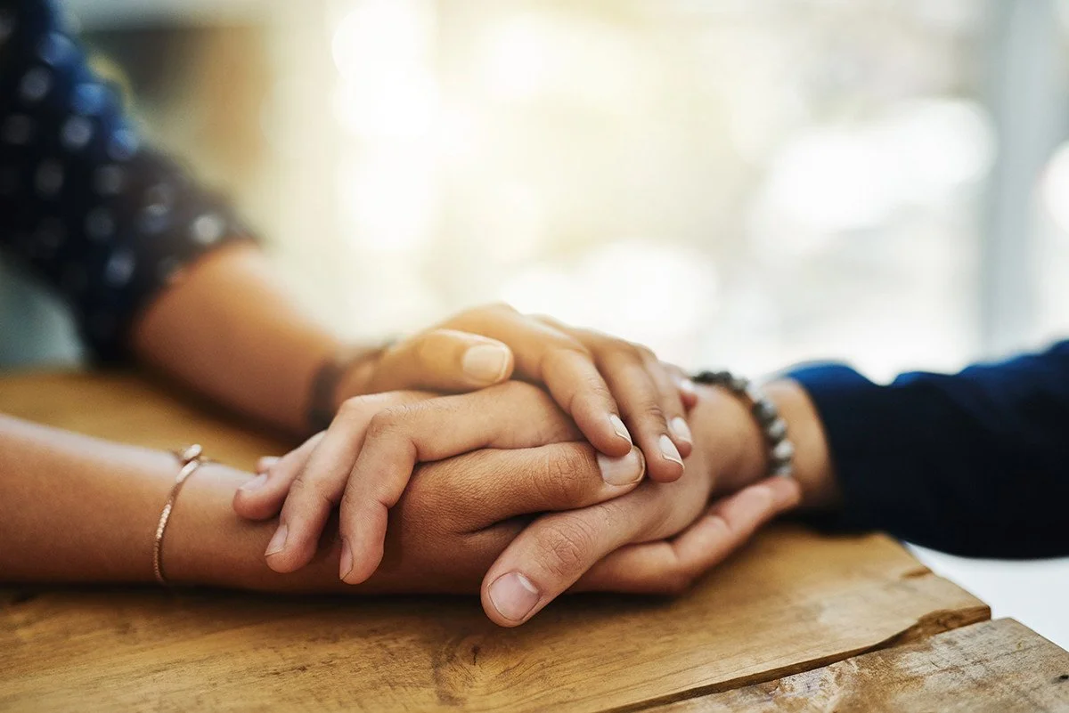 Two people holding hands on a wooden table, one person's hand gently resting on top of the other's.