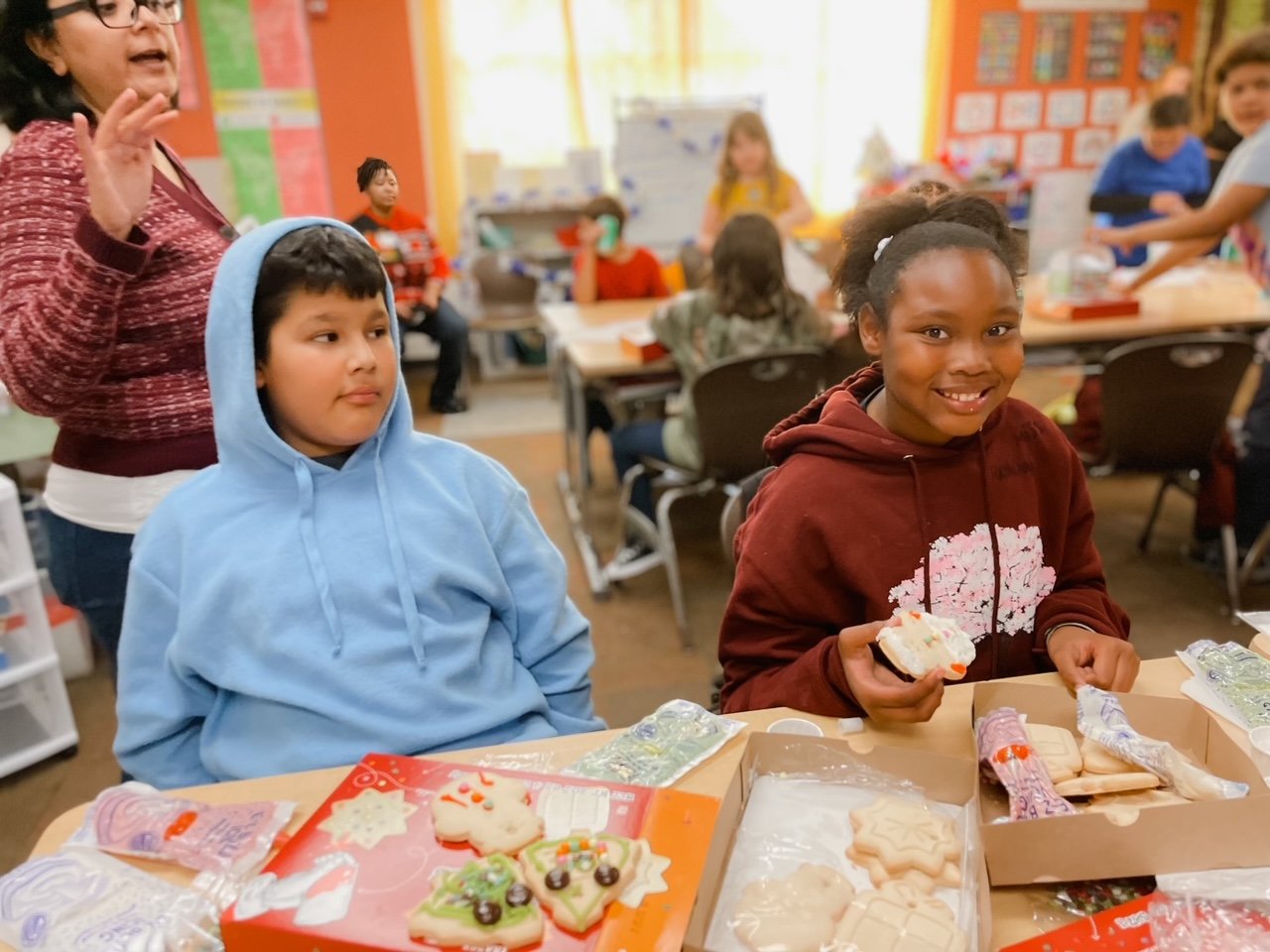Children celebrating at a classroom holiday party; two girls, one in a blue hoodie and one in a brown hoodie, smiling and holding decorated cookies; cookies in festive shapes and colors on the table.