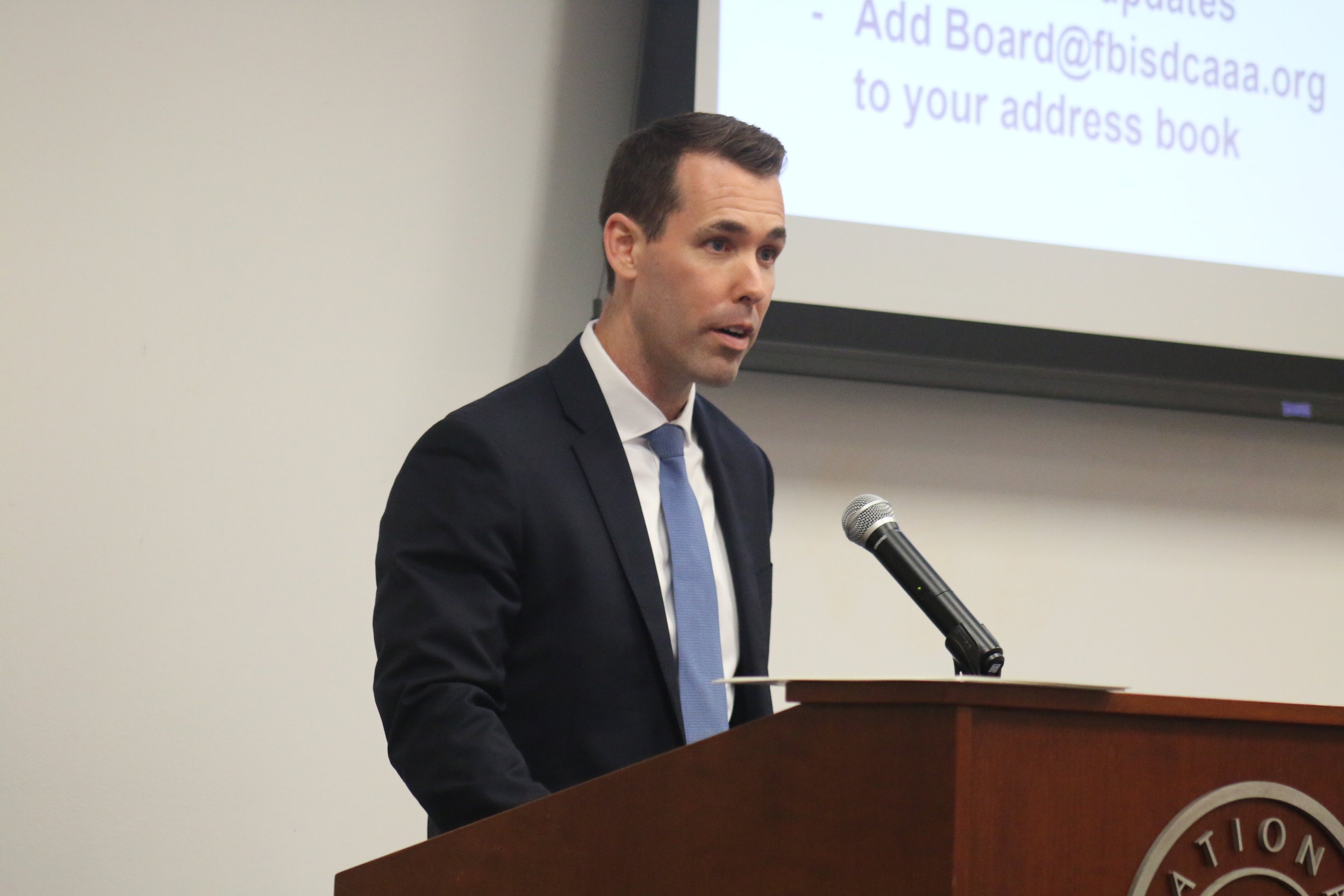 A man in a dark suit, white shirt, and blue tie speaking at a podium with a microphone, with a presentation slide projected behind him.