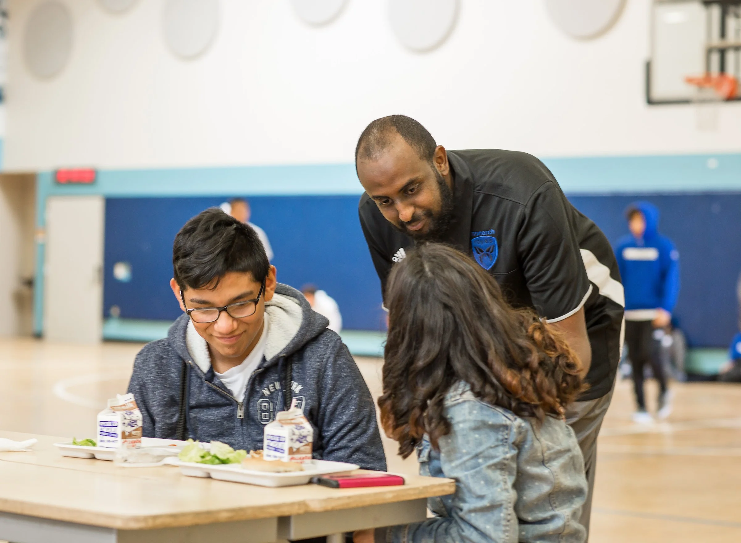 A coach interacts with two children sitting at a table during a meal in a gymnasium.