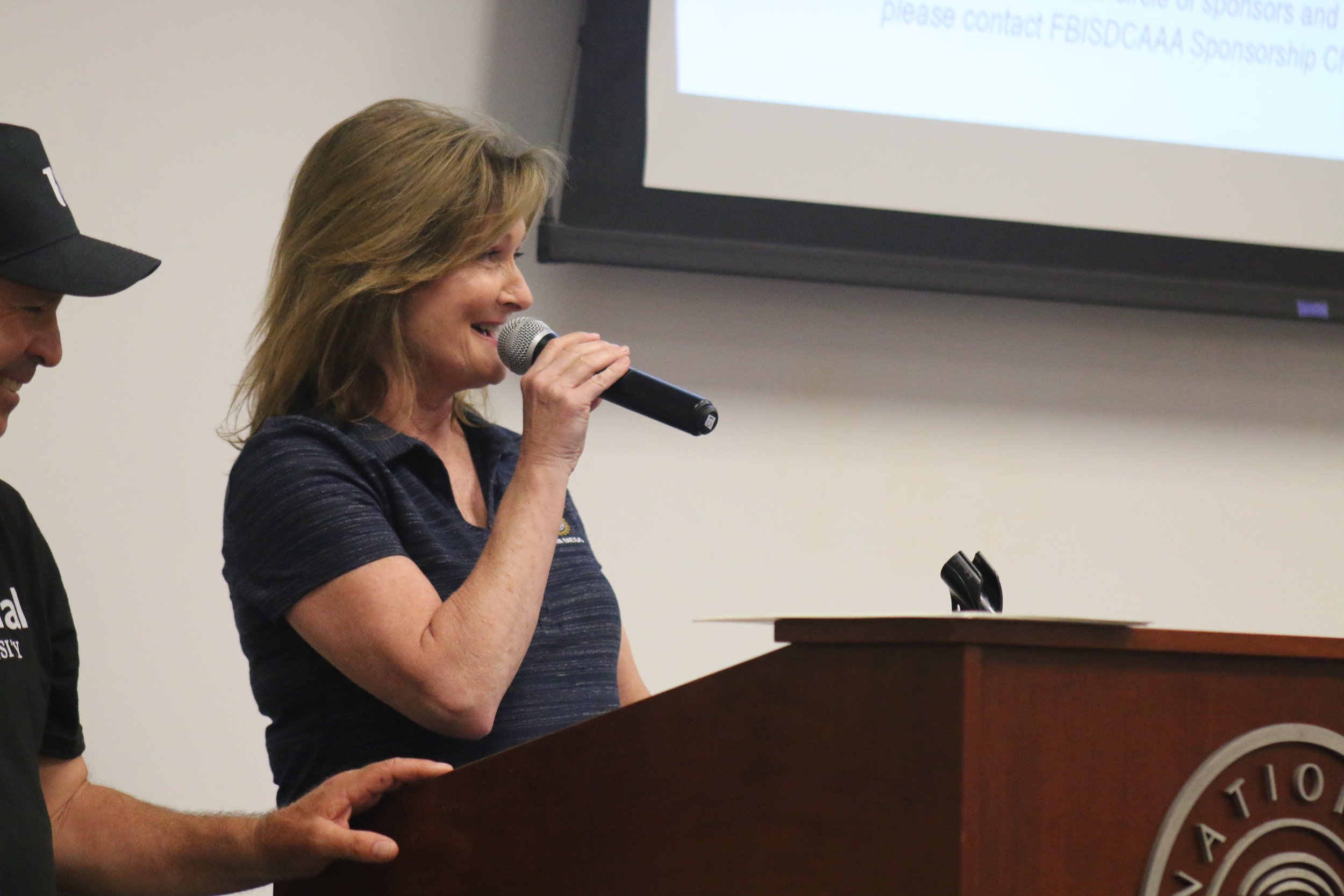 A woman with shoulder-length hair speaking into a microphone at a podium, with a screen displaying a presentation behind her.