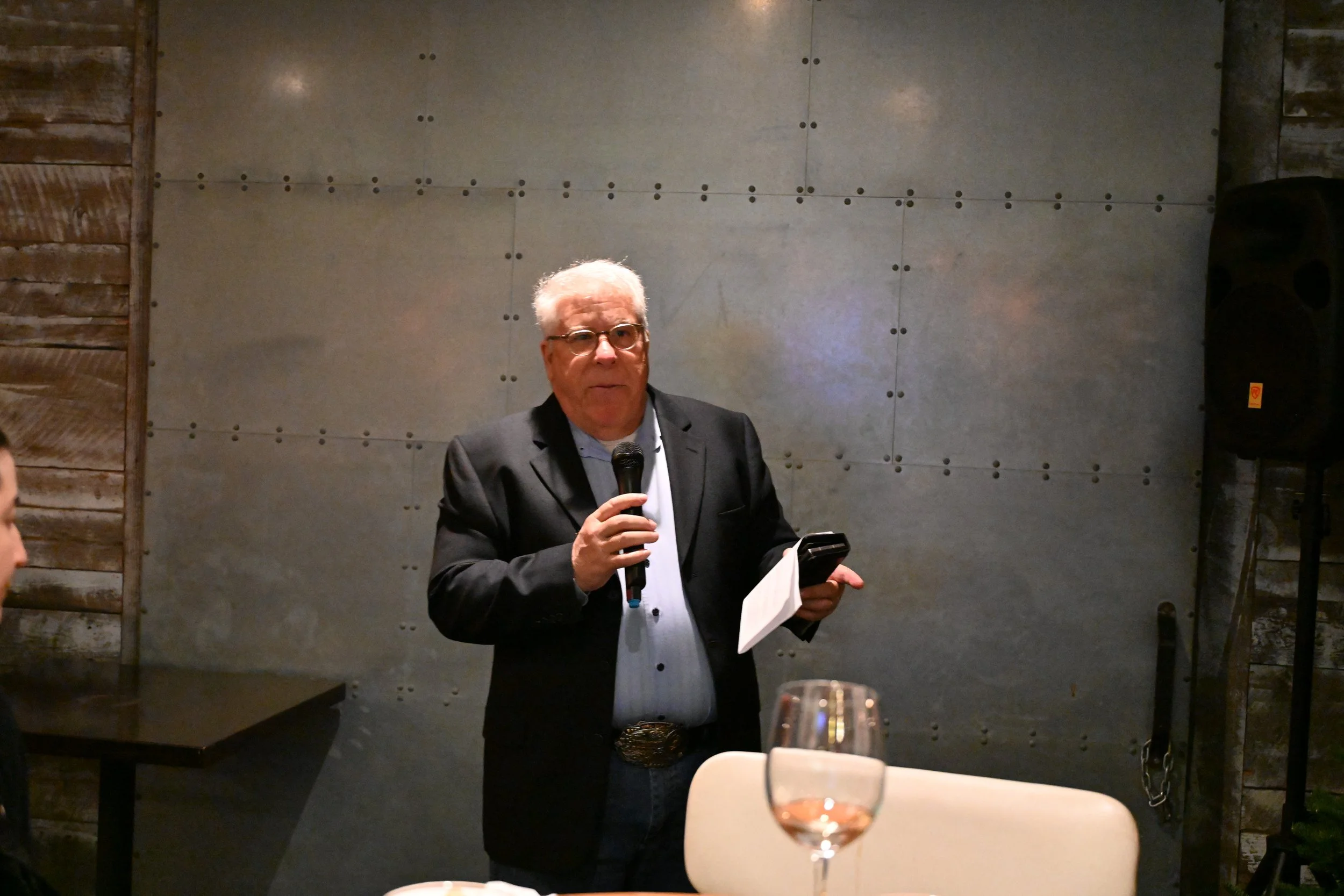 An older man with white hair and glasses wearing a black blazer, standing at a table, holding a microphone in one hand and a phone and paper in the other, speaking in front of a metallic wall in a dimly lit room.