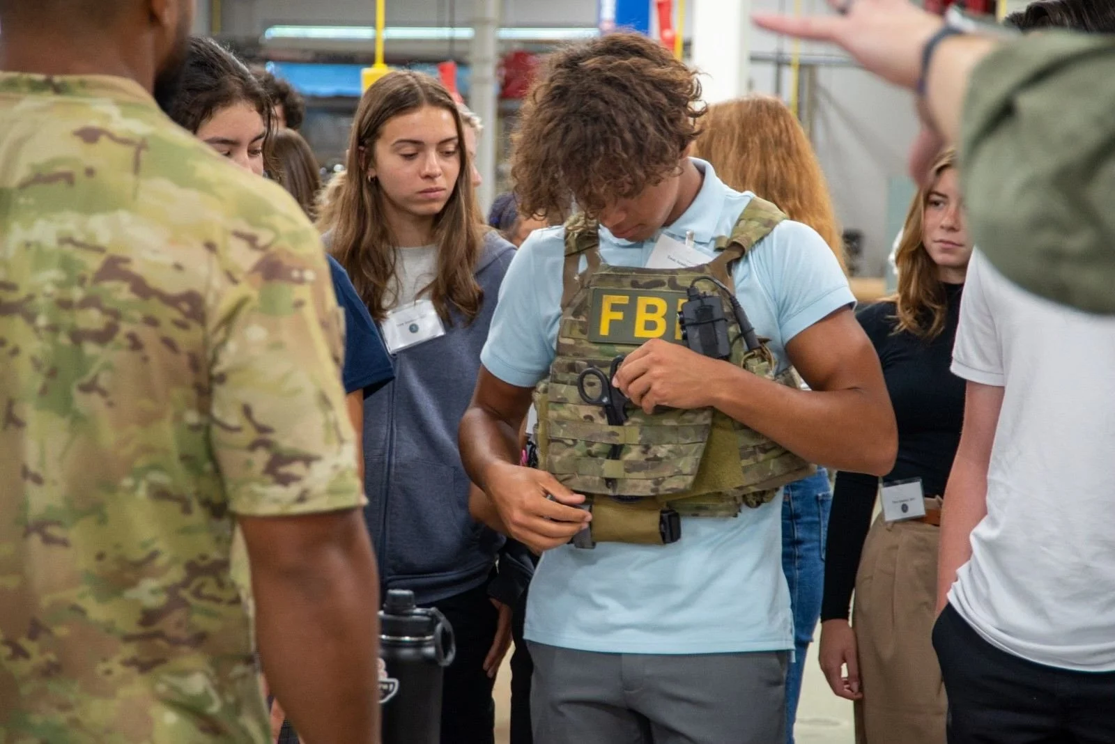 Group of young people gathered around a person in a light blue shirt and tactical vest with 'FBI' patches, inside a warehouse or industrial setting.