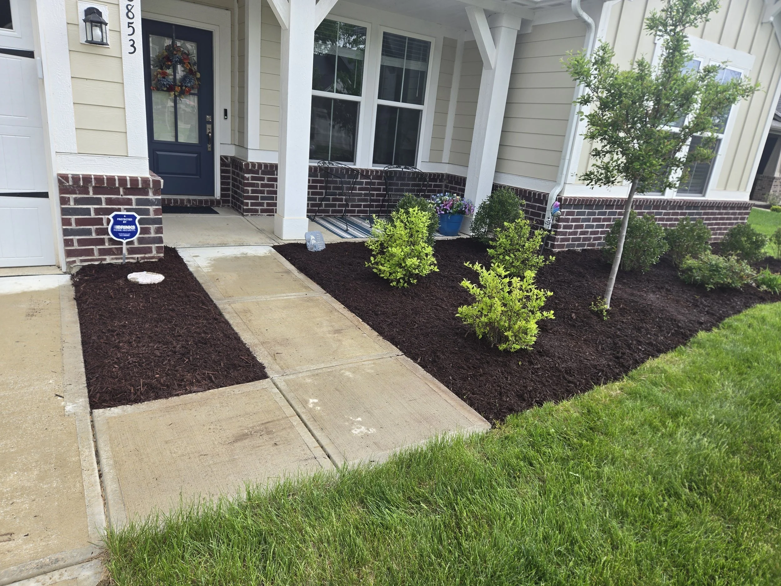 A front yard garden bed with mulch, small green bushes, and a decorative sunflower stake. In the background, there's a porch with outdoor chairs, a small table, and a security sign.