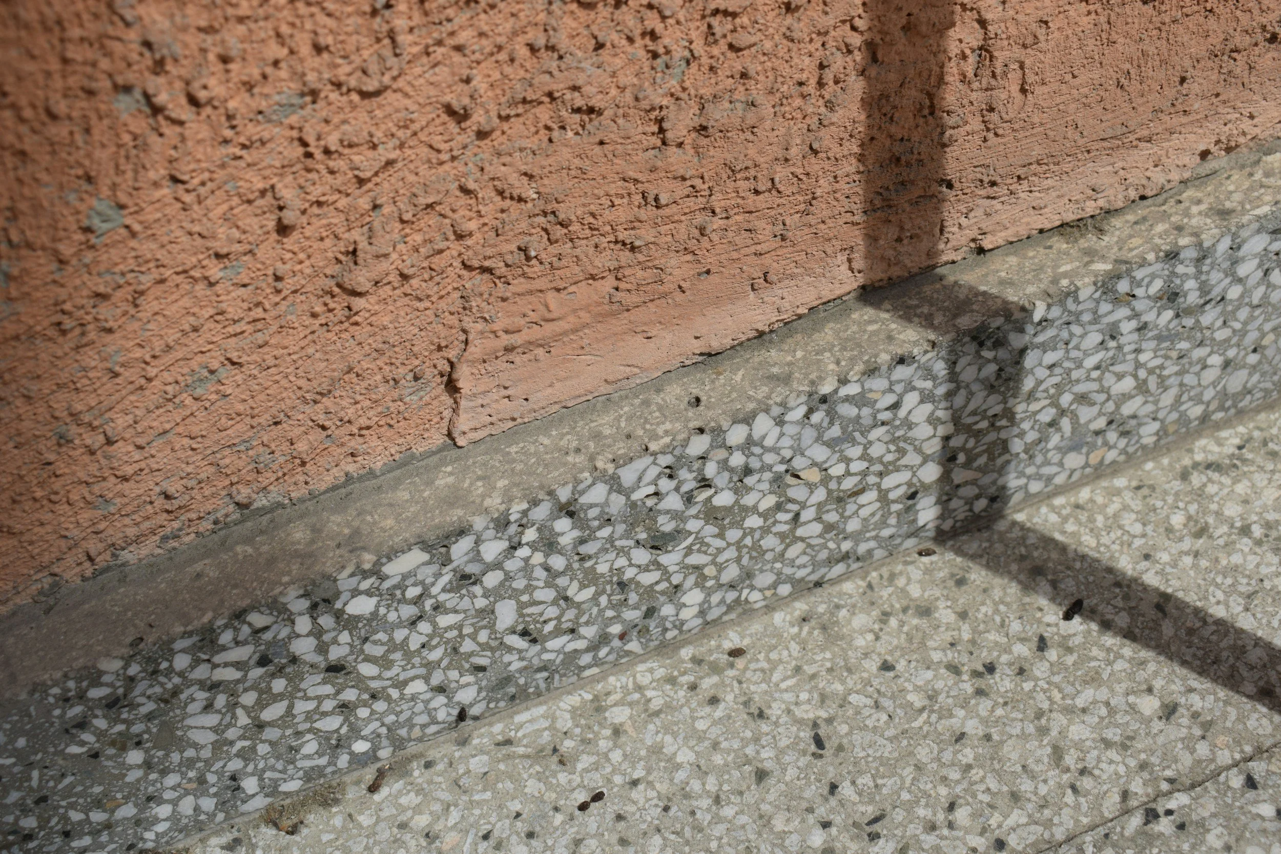 Close-up of a textured brick wall with a shadow cast on the ground, which is covered with small stones, in sunlight.
