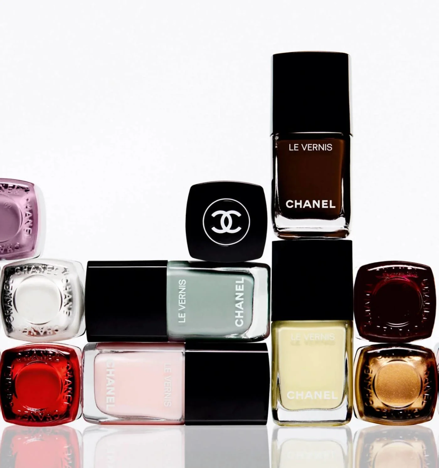 A collection of Chanel nail polish bottles in various colors, arranged with some bottles upright and others lying on their sides, on a reflective surface.