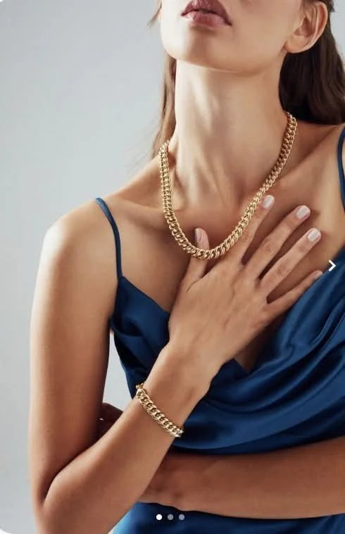 A woman wearing a blue satin dress, gold chain necklace, and matching bracelet, with her hand on her chest.