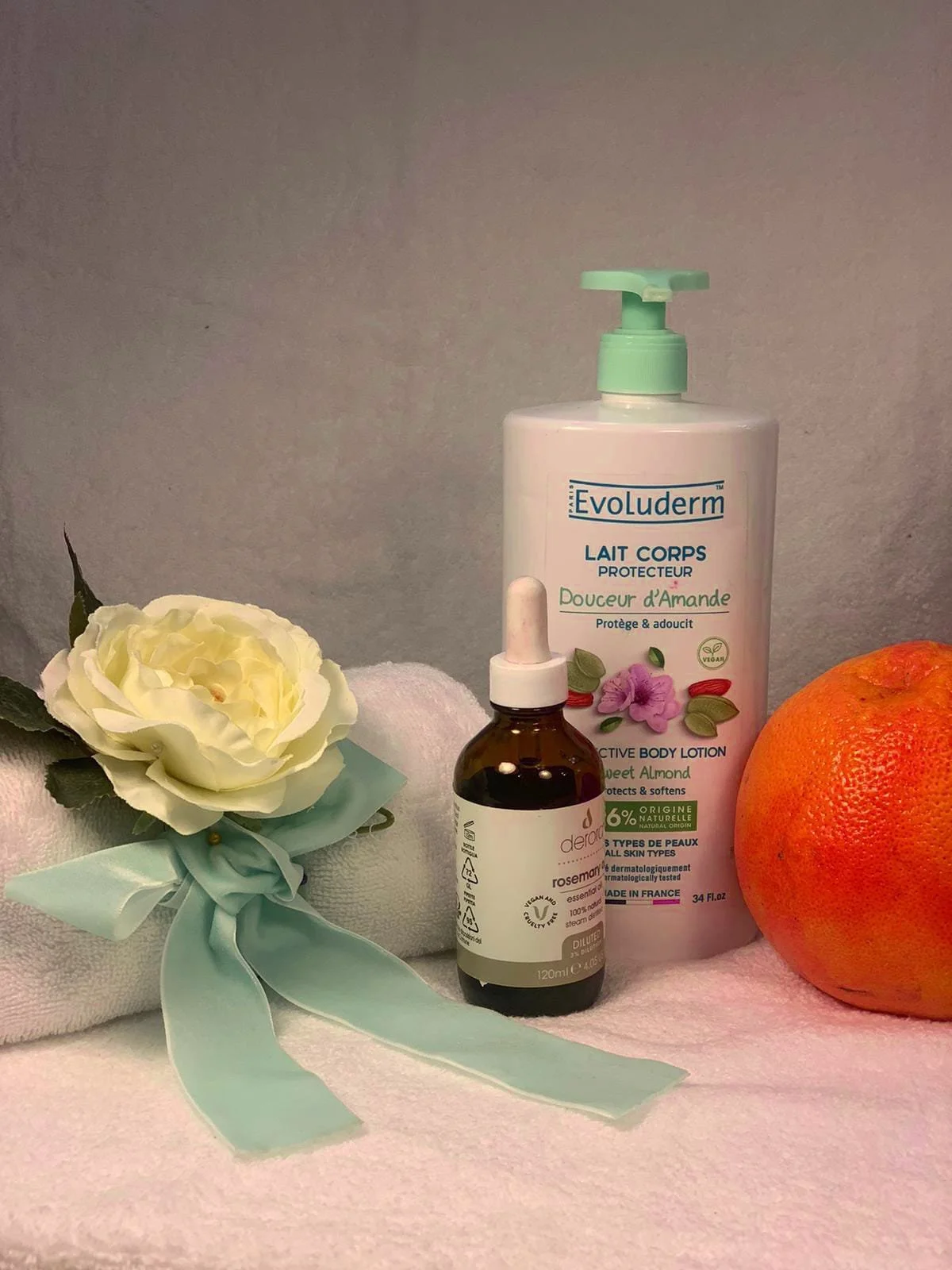 A flat lay of a white towel with a white flower with green leaves, a bottle of Evoluderm body lotion, a small dropper bottle of rosehip essential oil, and a pink grapefruit.