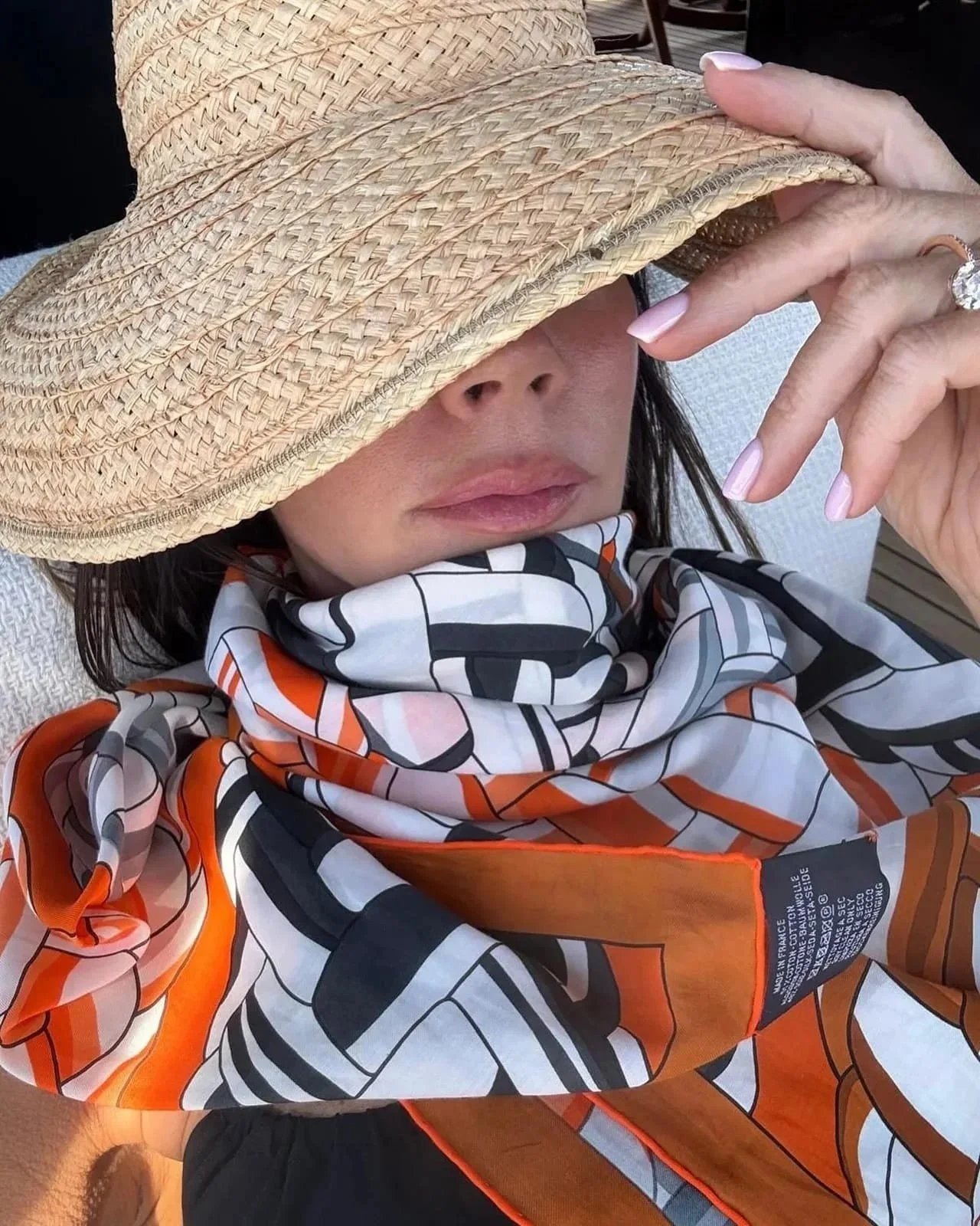 A woman wearing a wide-brimmed straw hat, a colorful patterned scarf, and a black top is sitting outdoors. She is holding the brim of her hat with her left hand, partially covering her face, showing only her lips and nose.