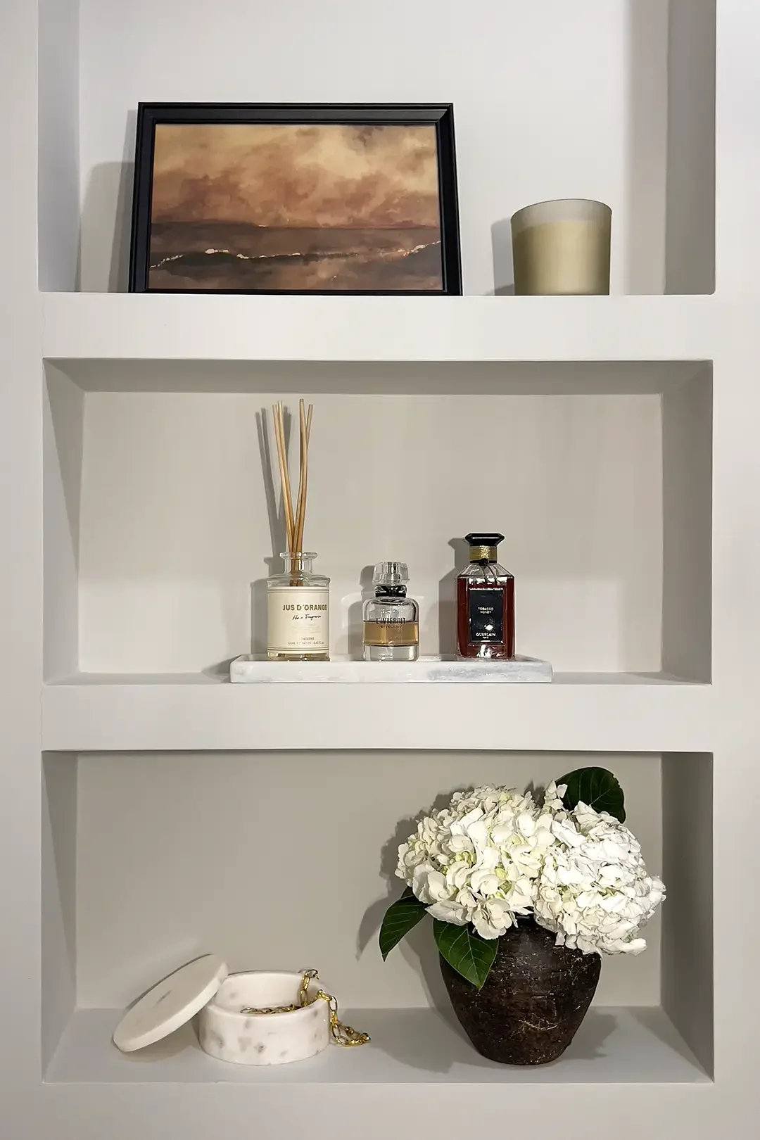 Built-in wall niche styled with framed artwork, candles, perfume bottles and vase of white hydrangeas by Studio Sakaia.”