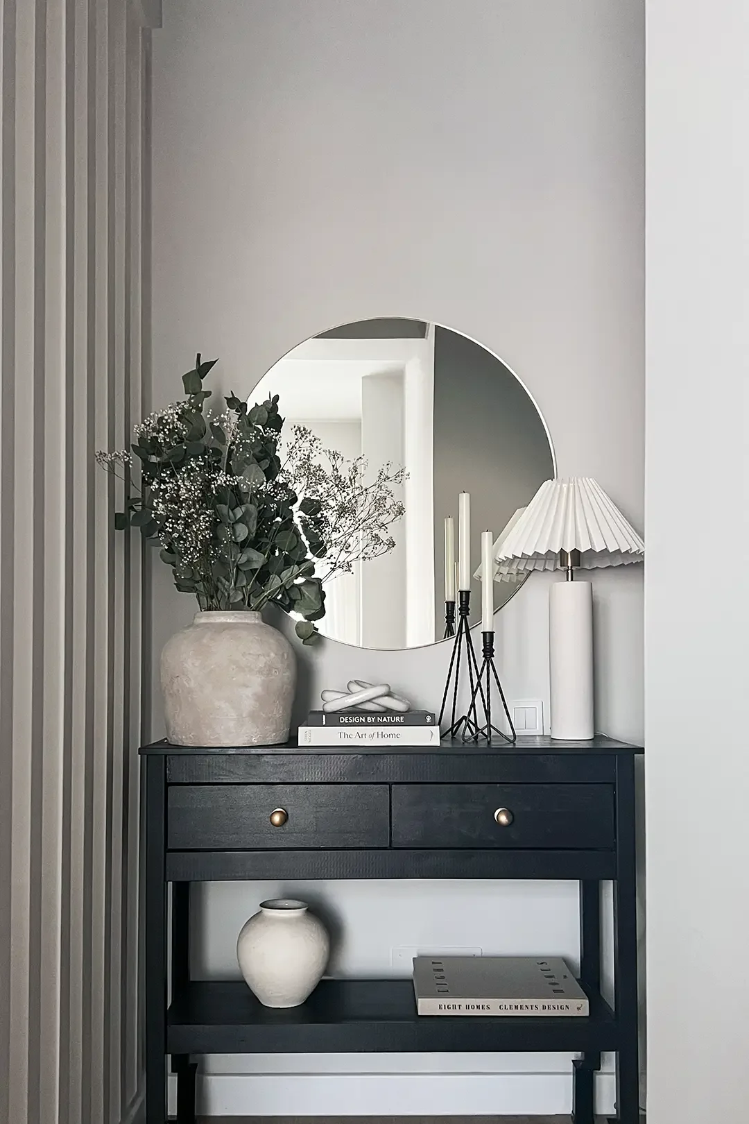 Styled black console by Studio Sakaia with greenery, round wall mirror, pleated table lamp and curated books.