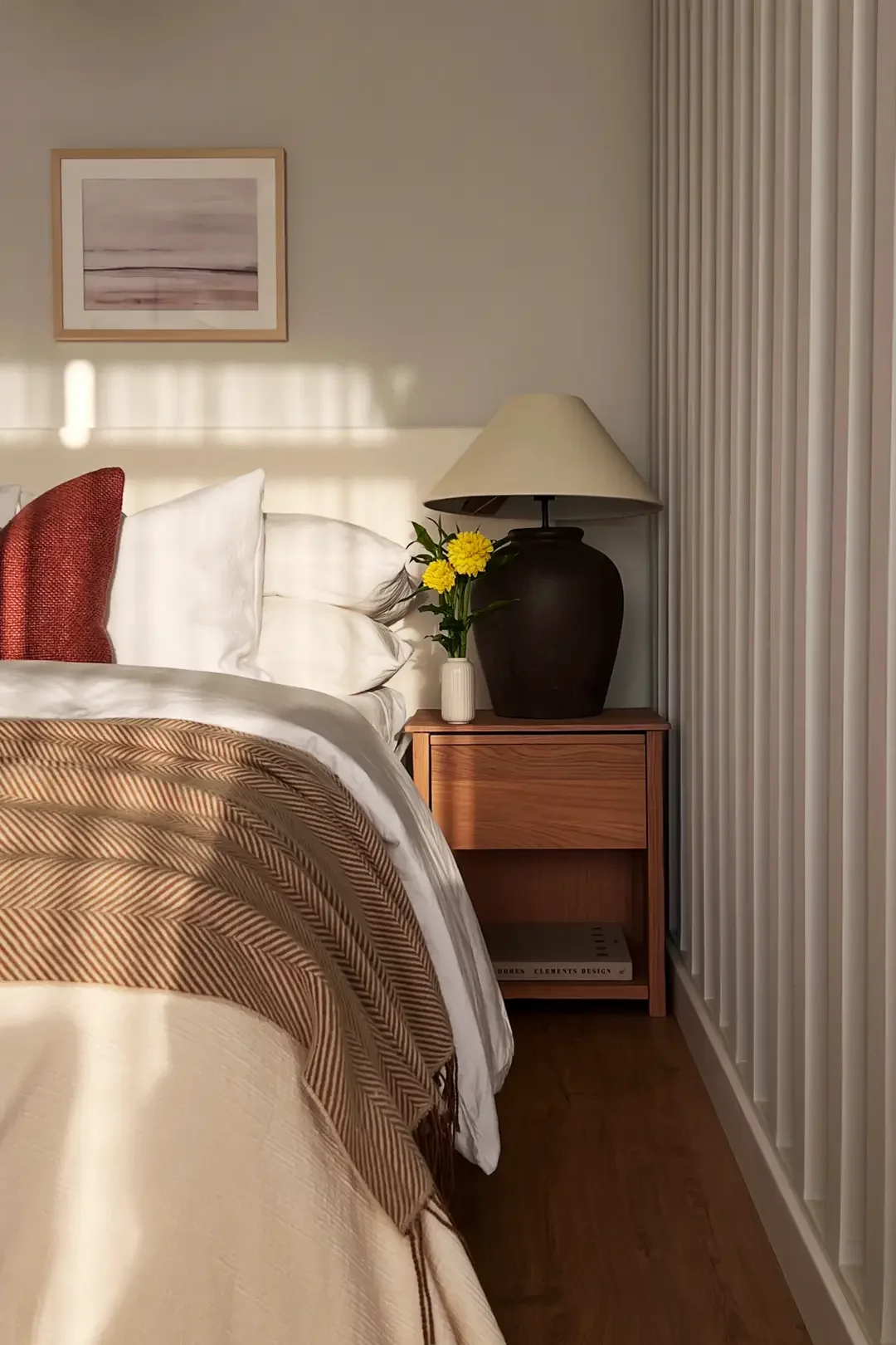 Bedroom corner styled by Studio Sakaia with layered white linens, rust accent pillow, striped throw, and wooden nightstand.