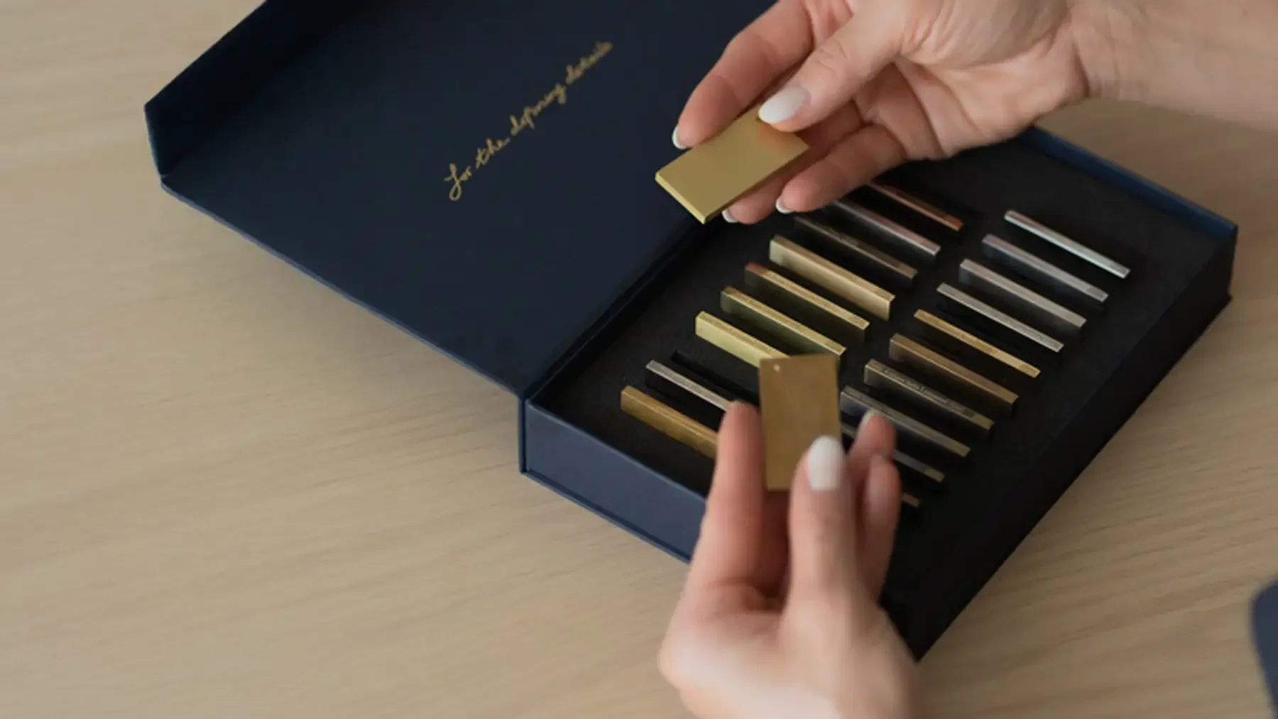 Madalina Aris selecting metallic finish samples from Studio Sakaia’s material presentation box on a pale oak surface.