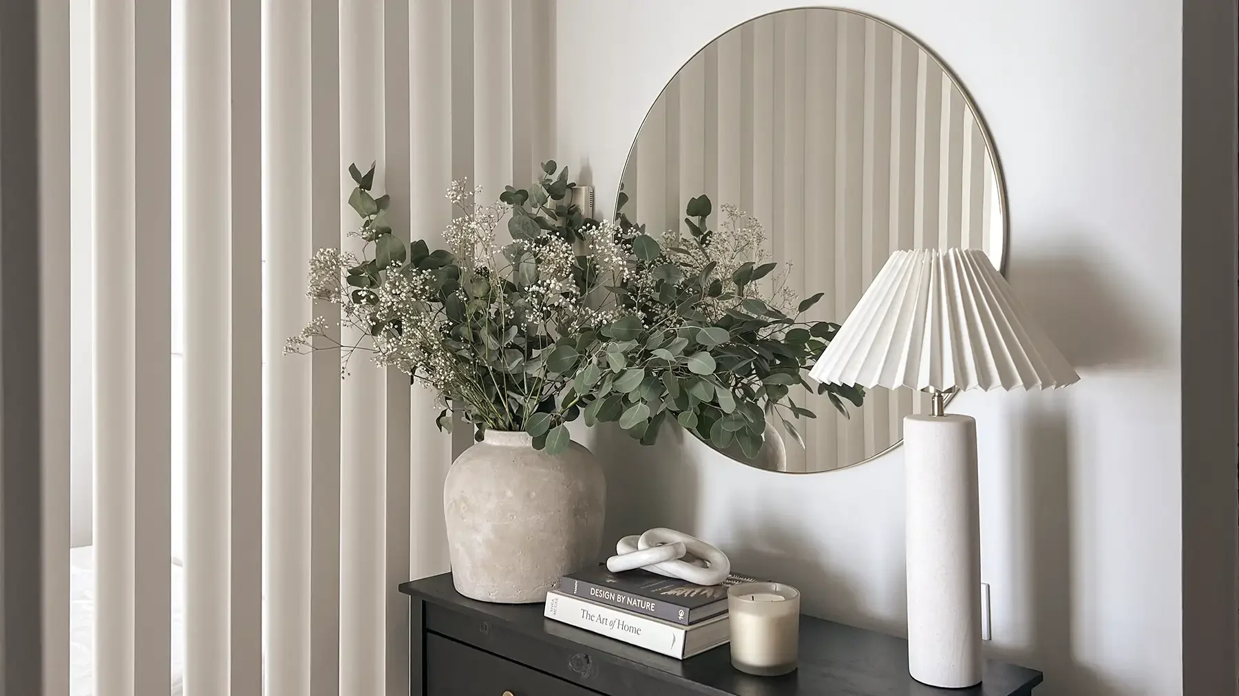 Styled corner with ceramic vase and greenery, sculptural knot, books and candle on a black side table, against vertical white panels, by Studio Sakaia.