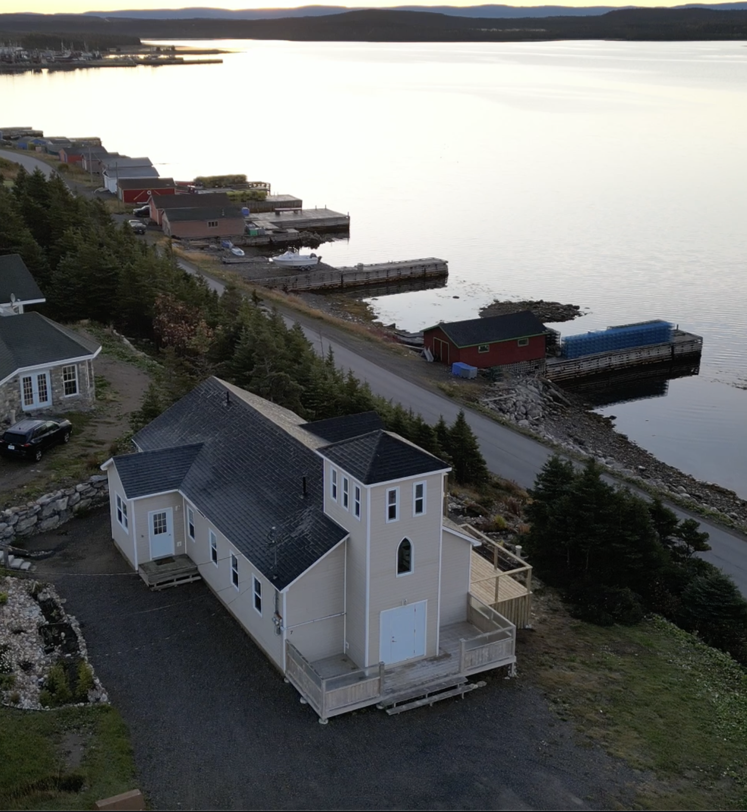 Aerial view of a coastal neighborhood with houses, including a prominent white church-style home in the foreground, and boats docked along the shoreline under an evening sky.