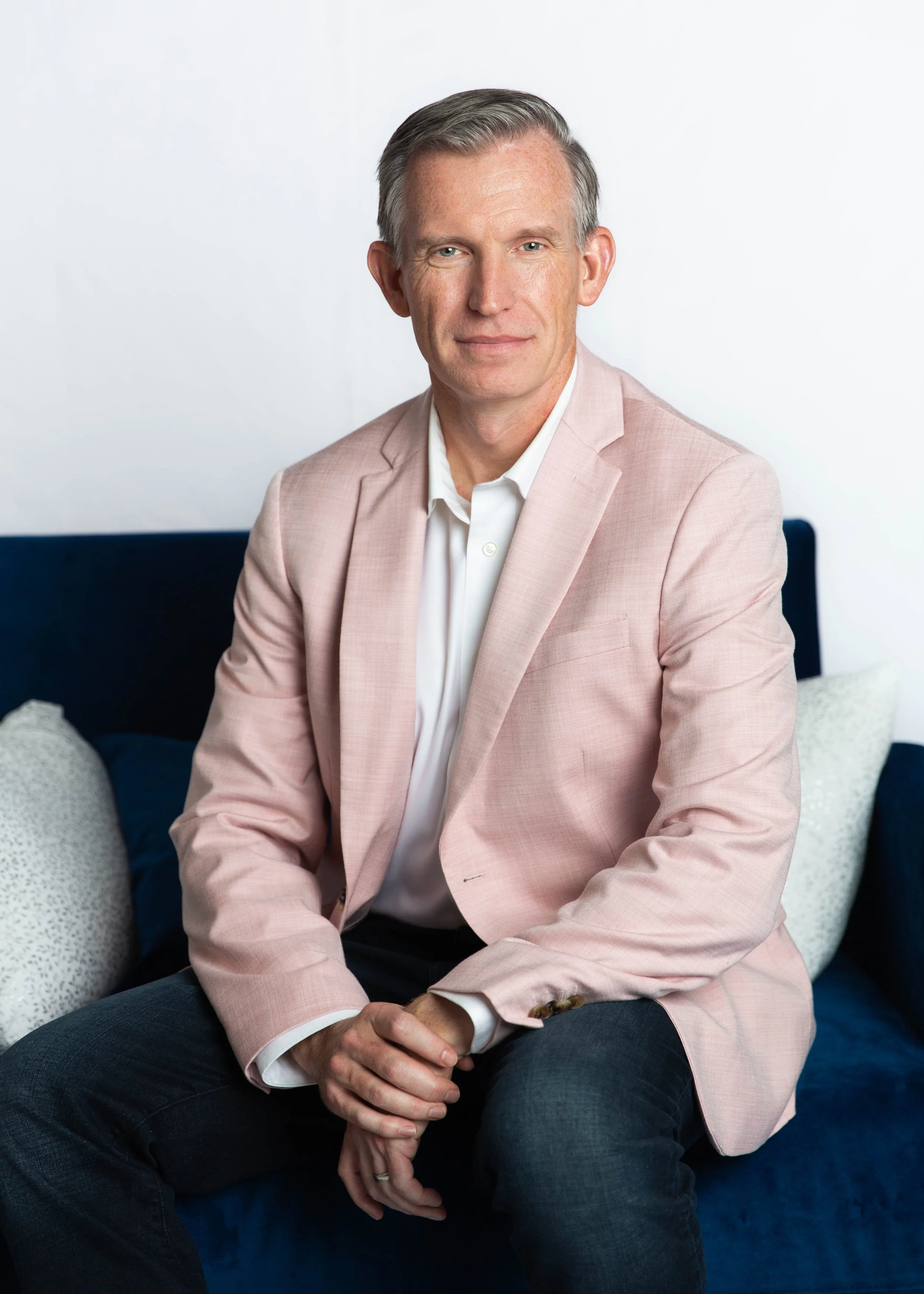 Brian L. Reece in a light pink blazer and white shirt sitting on a navy blue couch with decorative pillows, against a plain white background.