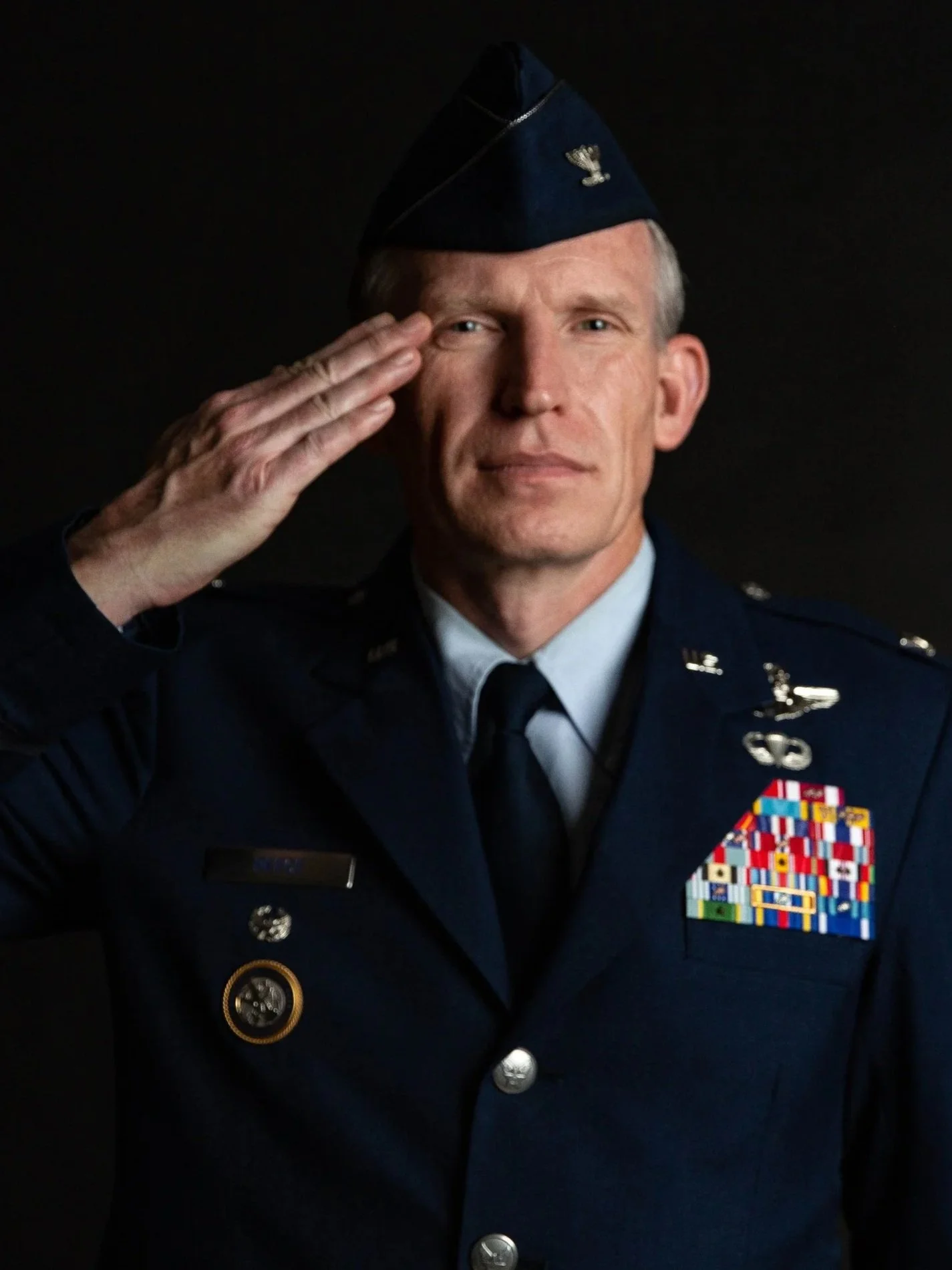 Colonel Brian L. Reece in a United States Air Force uniform salutes, wearing a blue service cap and decorated with medals and ribbons on his chest.