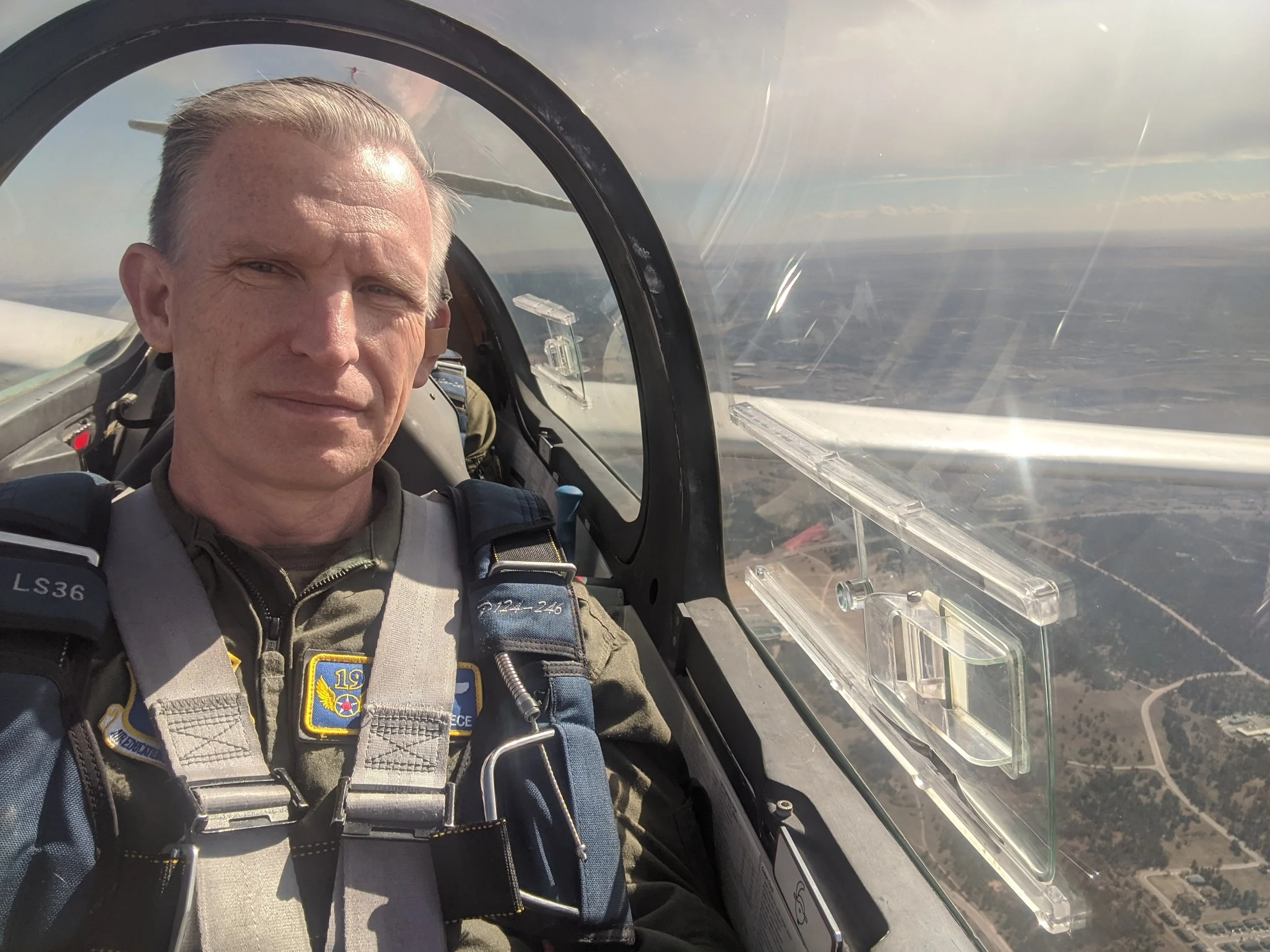 Brian Reece pilot gear inside the cockpit of a glider, with a view of the sky and landscape below through the cockpit window.