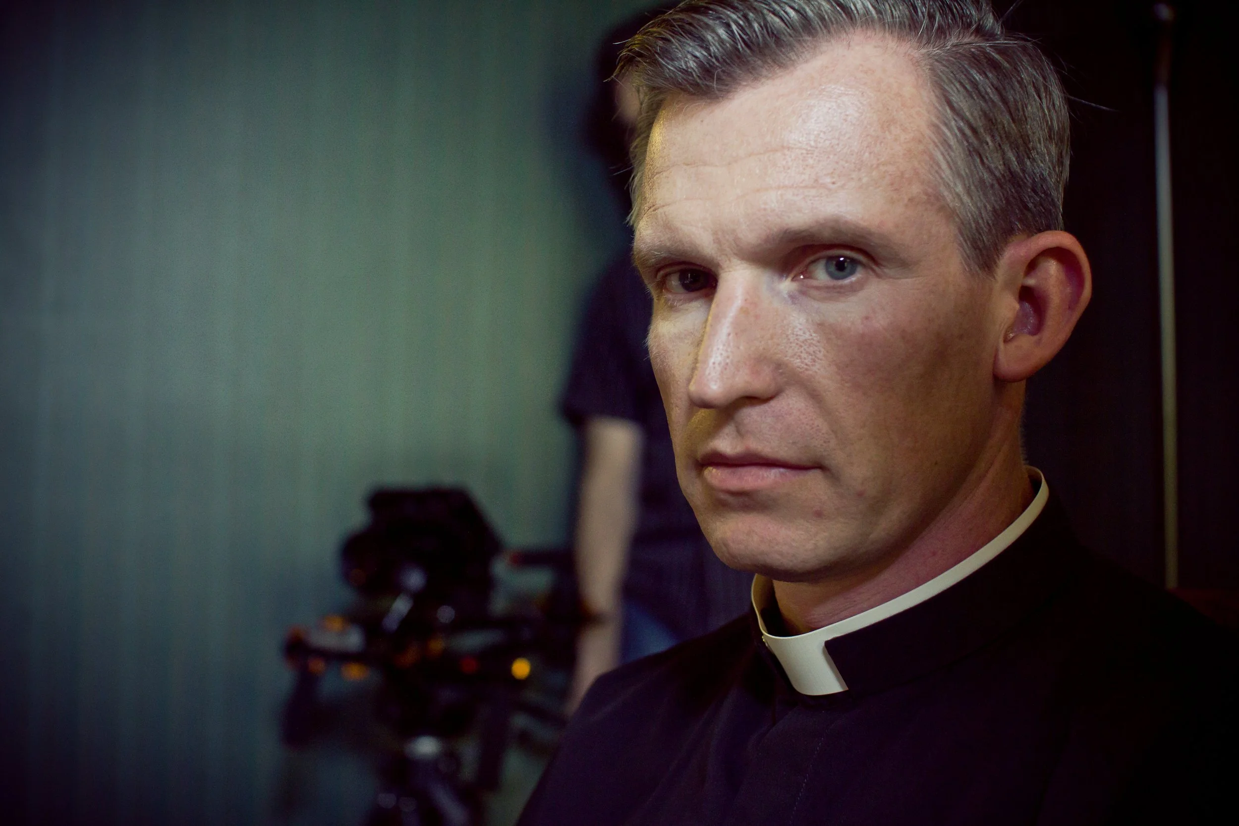 Close-up of a Brian Reece playing a priest on Blindsight. He has short, light-colored hair and blue eyes, wearing a black clerical shirt with a white collar, standing in front of a blurred background.