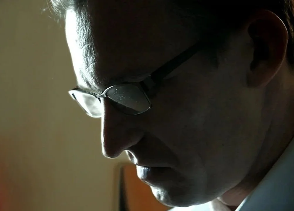 A close-up side profile of a Brian Reece wearing glasses, focused on his face and looking down, with subdued lighting highlighting the contours of their face.