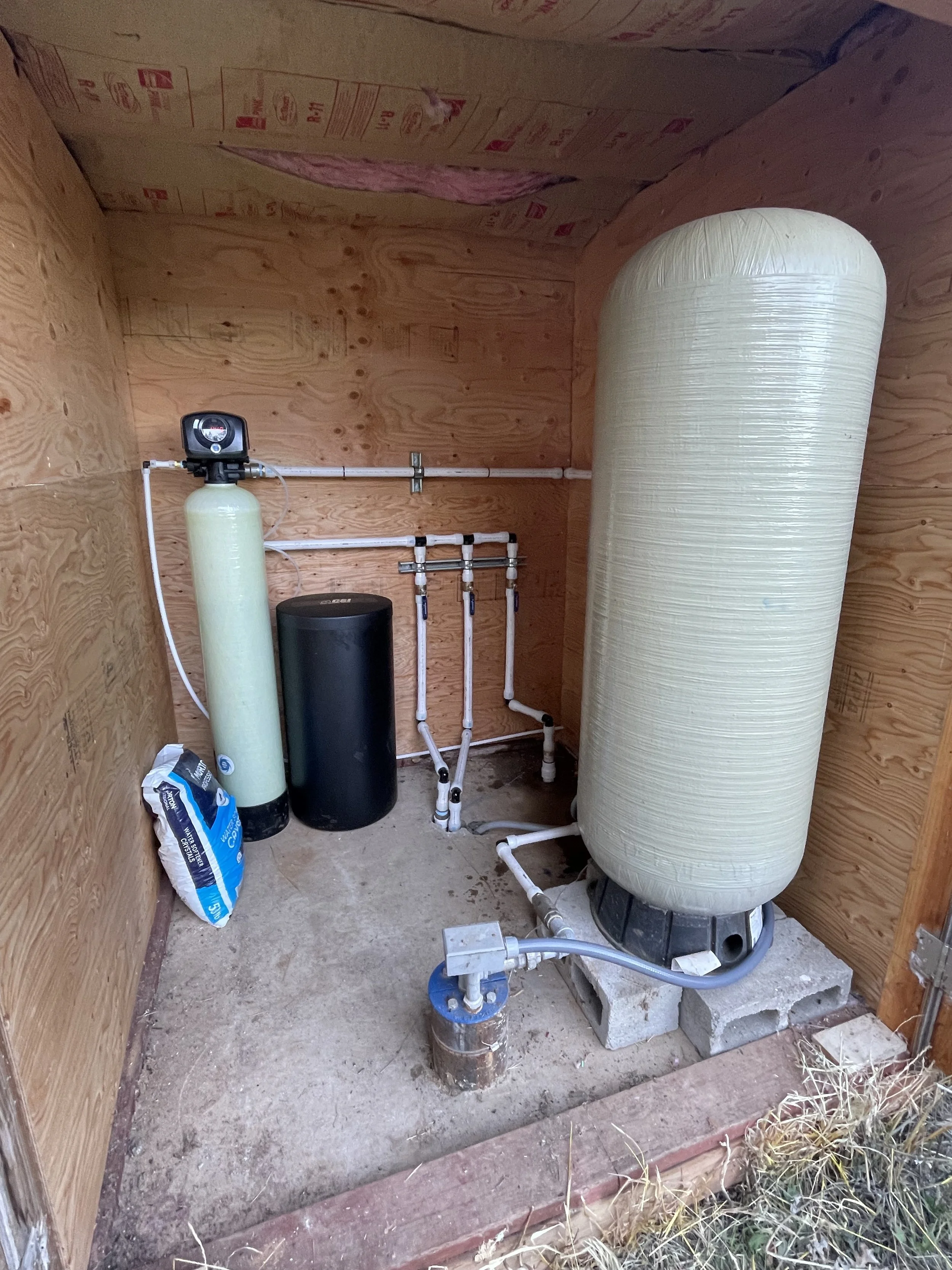 A small utility room with a large water filtration tank, a black sediment filter, and various pipes and fittings.
