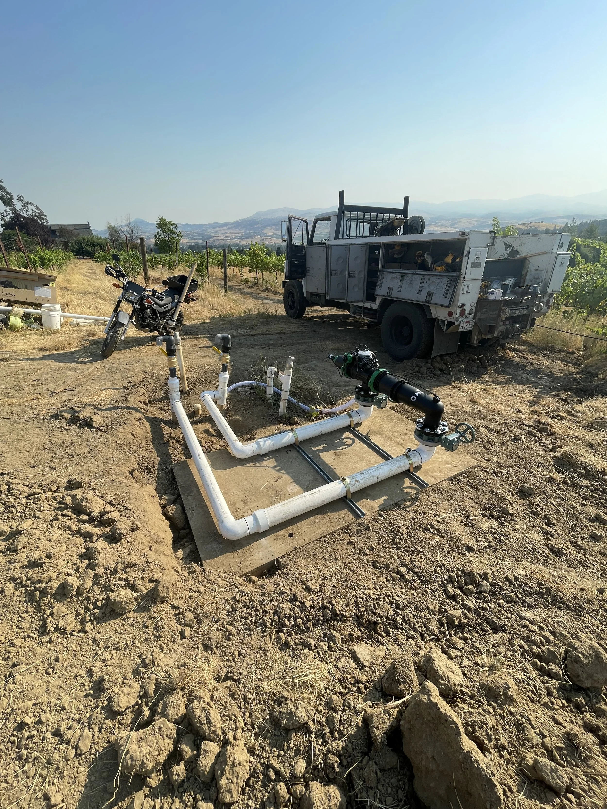 A irrigation water filter system installed on a concrete slab in a vineyard with a utility truck and motorcycle nearby, scenic hills in the background.