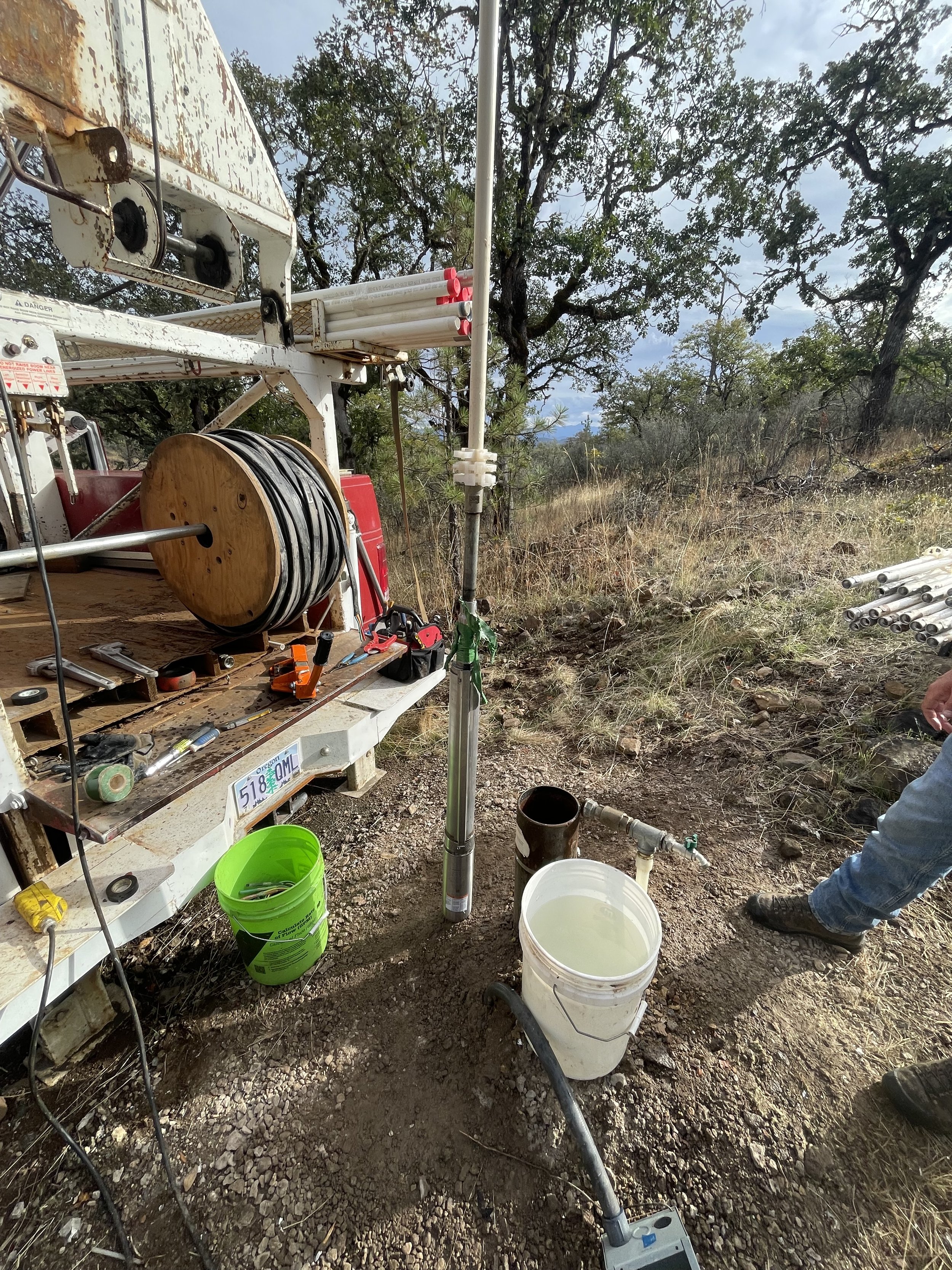 A water well installation setup outdoors with pipes, tools, and equipment on the ground and a bucket of water, amid dry grass and trees.