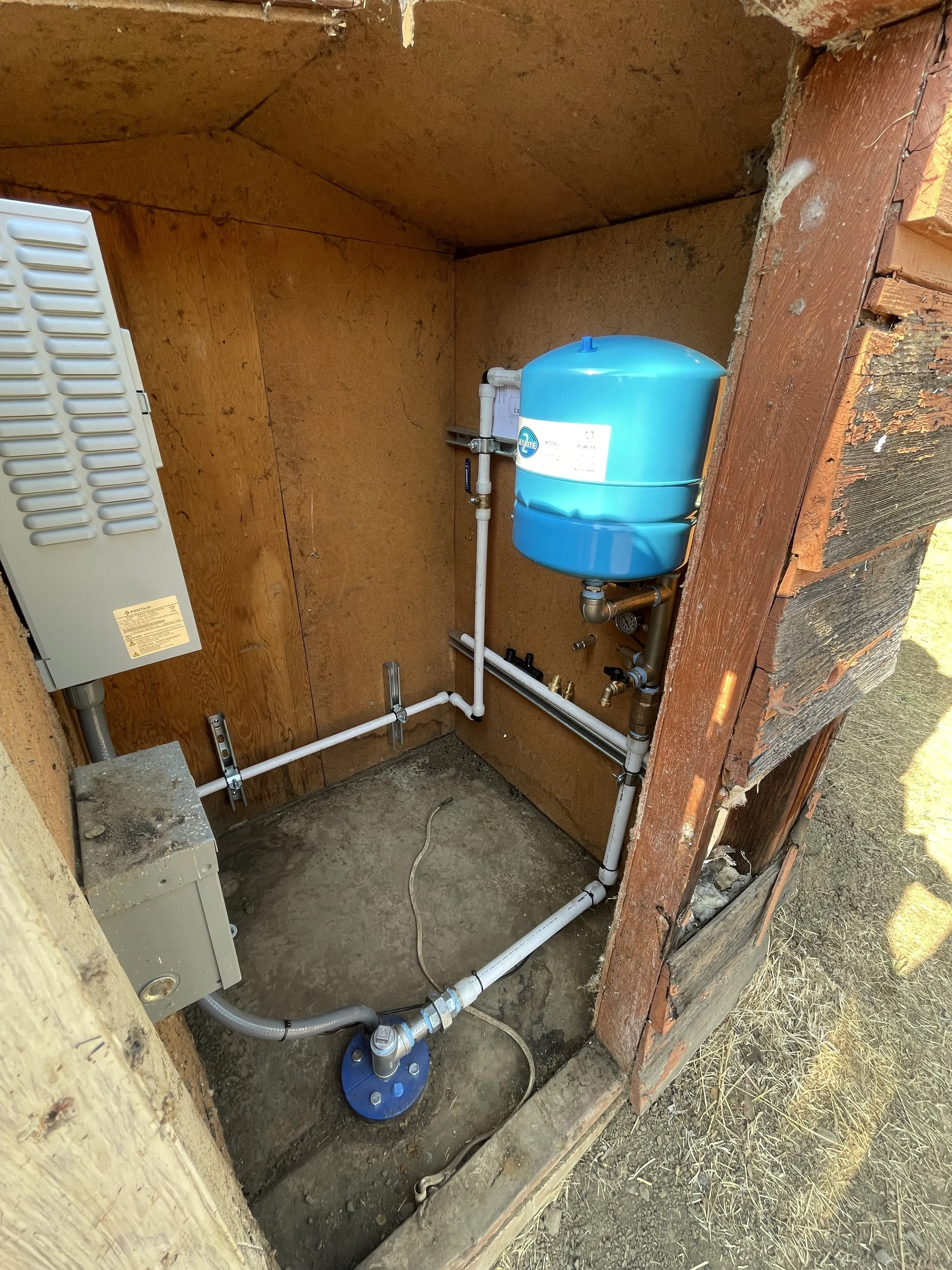 A well pump utility area with a blue pressure tank, plumbing pipes, an electrical box, and a gray ventilation unit inside a wooden enclosure with dirt floor.