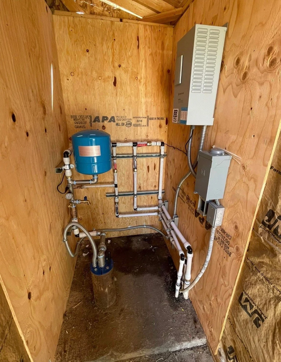 View of a well pump room with exposed plywood walls and floor, including pipes, gauges, a blue tank, and electrical boxes. a vfd drive unit and a water well