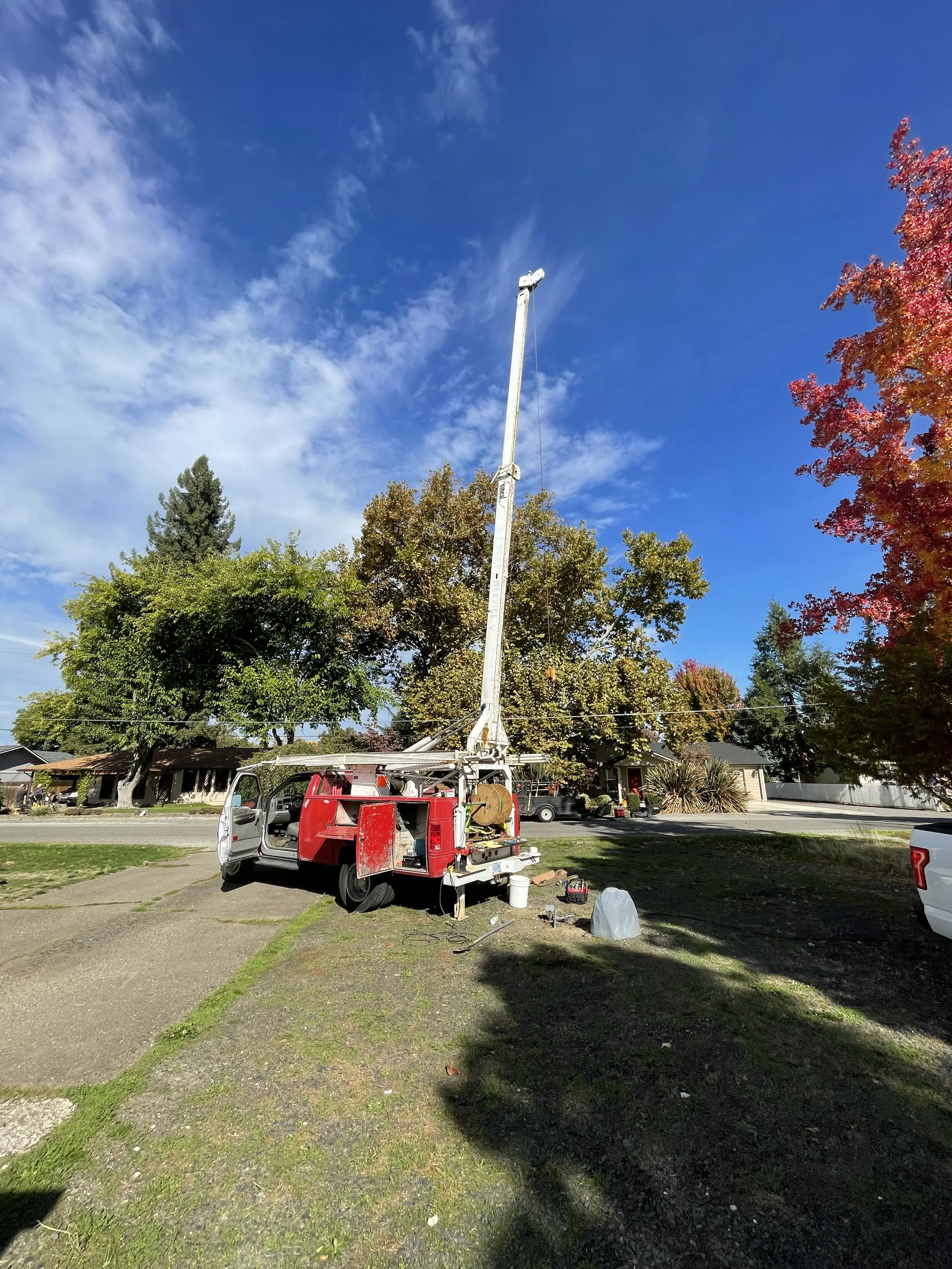 A well pump truck with a tall, extended crane working on a setting a well pump in a suburban neighborhood, with colorful fall foliage and clear blue sky.