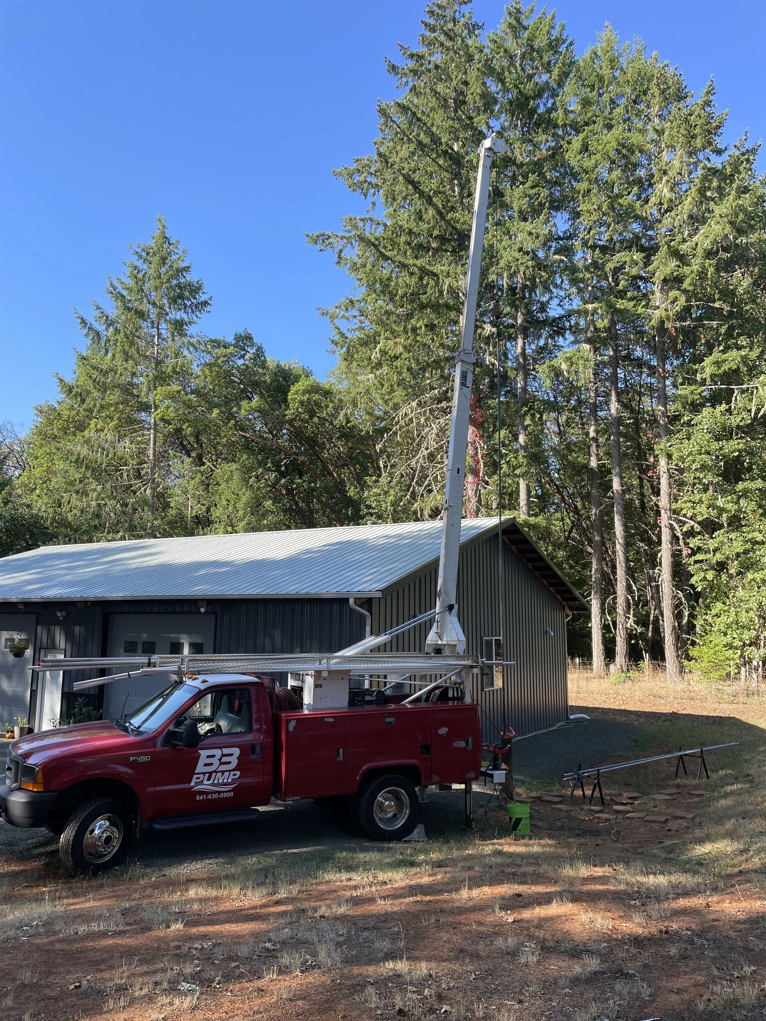 A red well pump hoist truck pulling a well pump is parked on a grassy area near a metal building, with forested trees in the background.