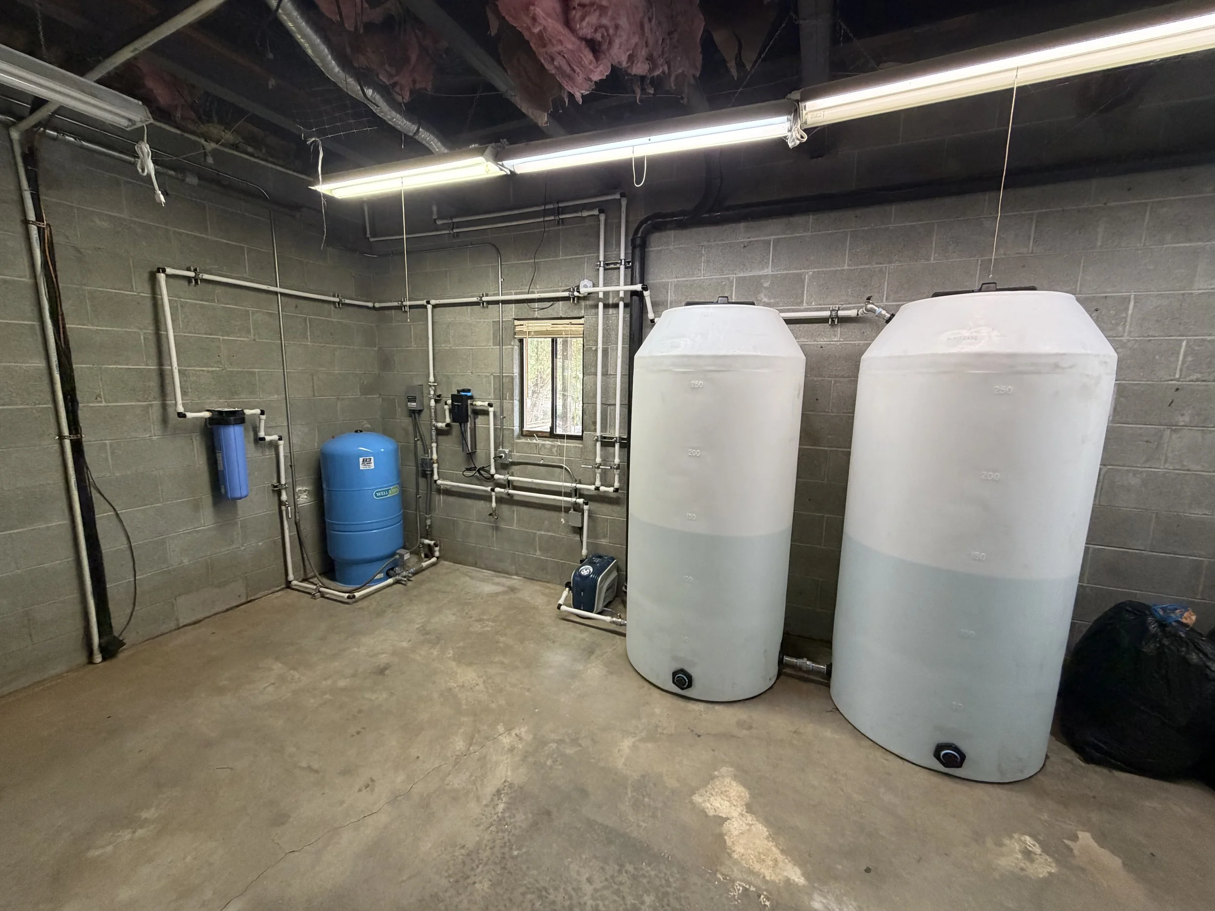 A utility room with water treatment equipment, including two large white tanks, a blue filter tank, and various pipes and controls on concrete walls and floor.