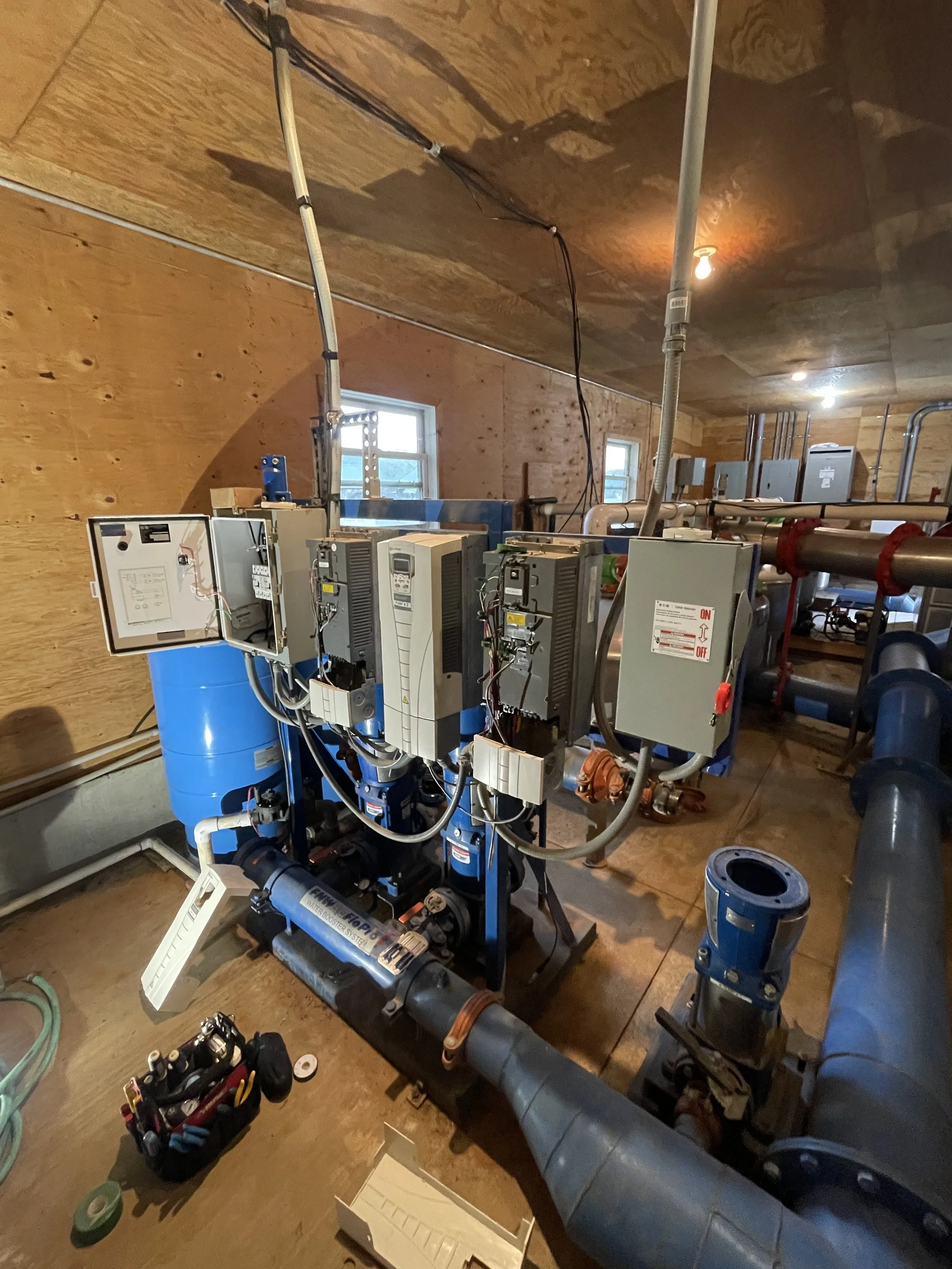 A mechanical irrigation room with various pumps, pipes, VFD's, electrical boxes, and wiring. The room has wooden walls, small windows, and is lit by ceiling lights.