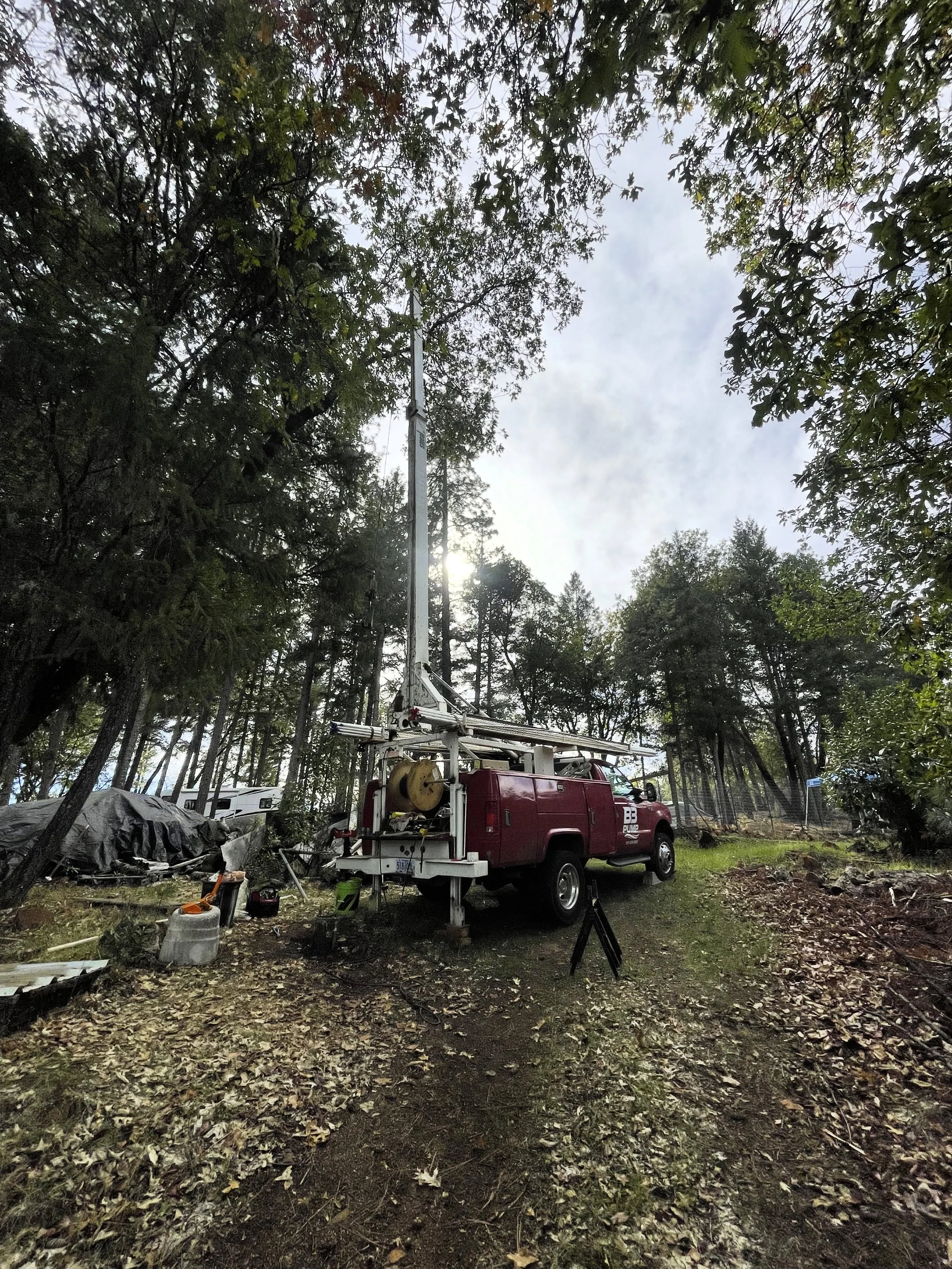 a well pump hoist truck with its crane up, situated in a wooded area with trees and fallen leaves.