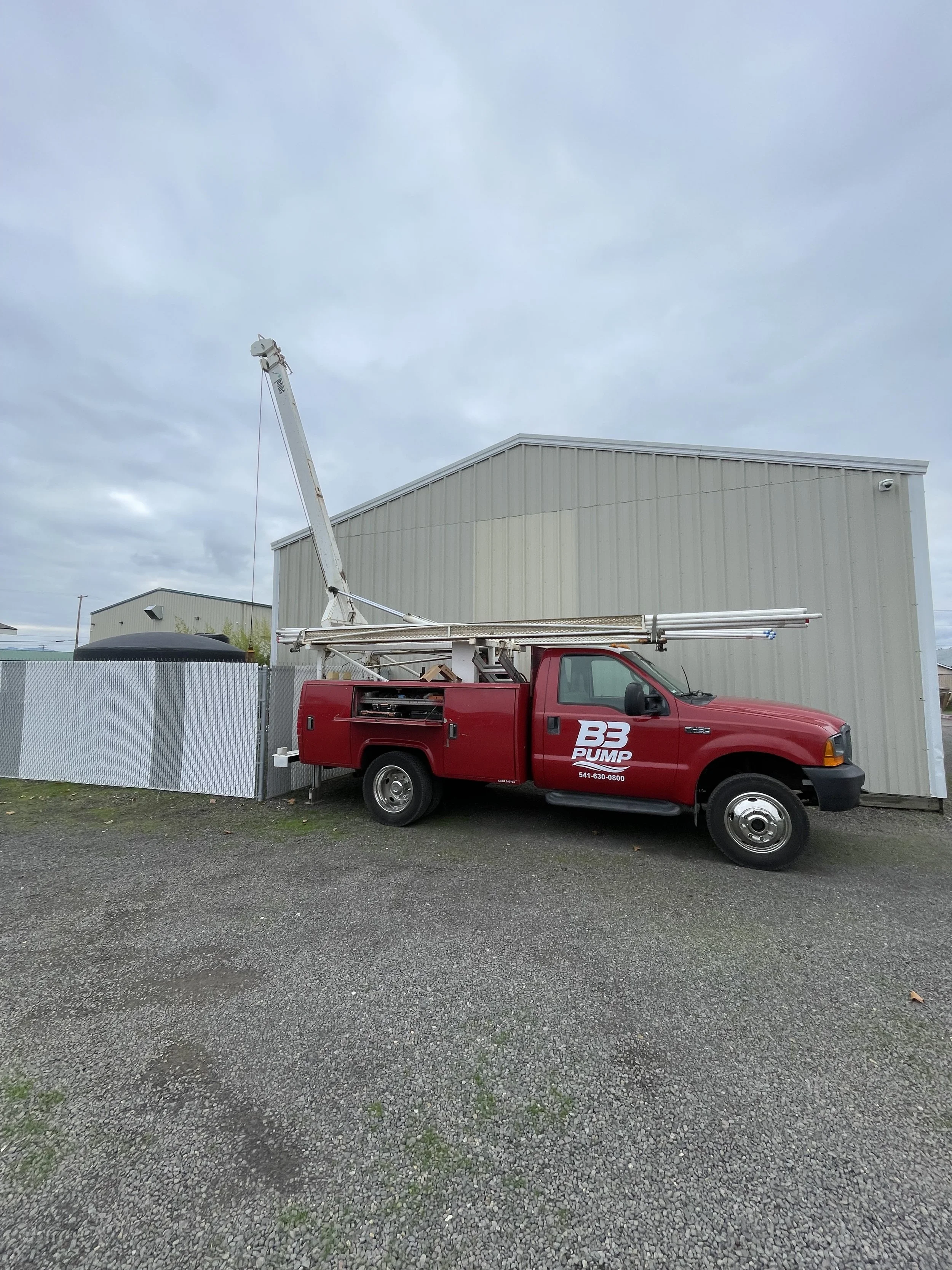 Red well pump hoist truck with a mounted ladder parked beside a beige metal building and a chain-link fence, overcast sky.