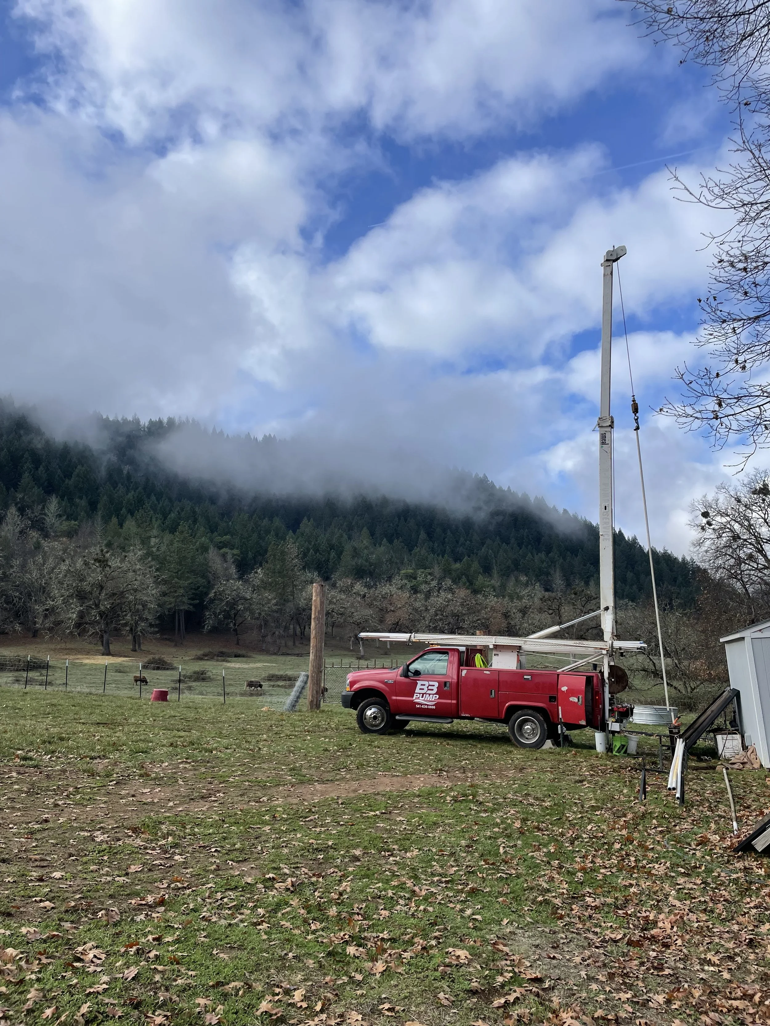 A red well pump truck with an extendable crane is parked and pulling a well pump on a grassy area near a fence with rolling hills and a forest in the background. The sky is partly cloudy.