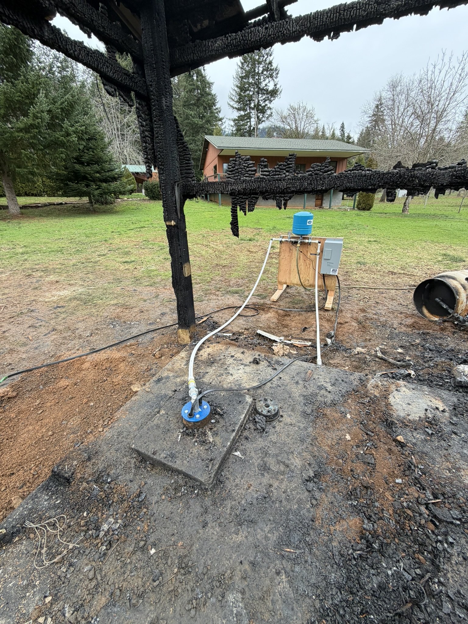 Burnt wooden structure with charred beams, electrical components and wires connected to a small blue water tank, on a dirt patch with grassy yard and trees in the background. temp well manifold set up in grants pass