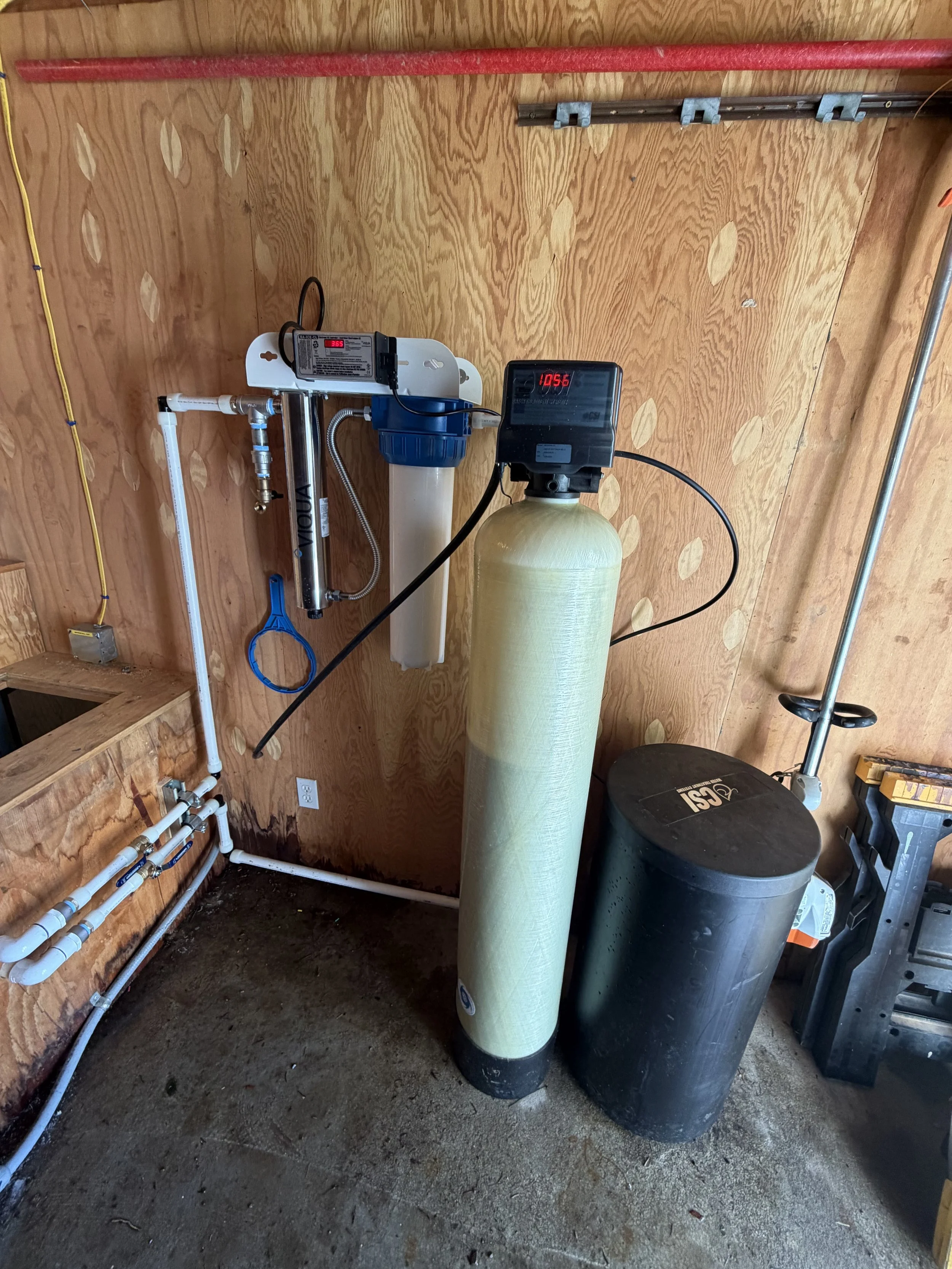 Garage with water filtration system, including a tall green water softener tank, a control valve with digital display, and black brine tanks. Pipes and electrical wiring are visible on the wooden wall.