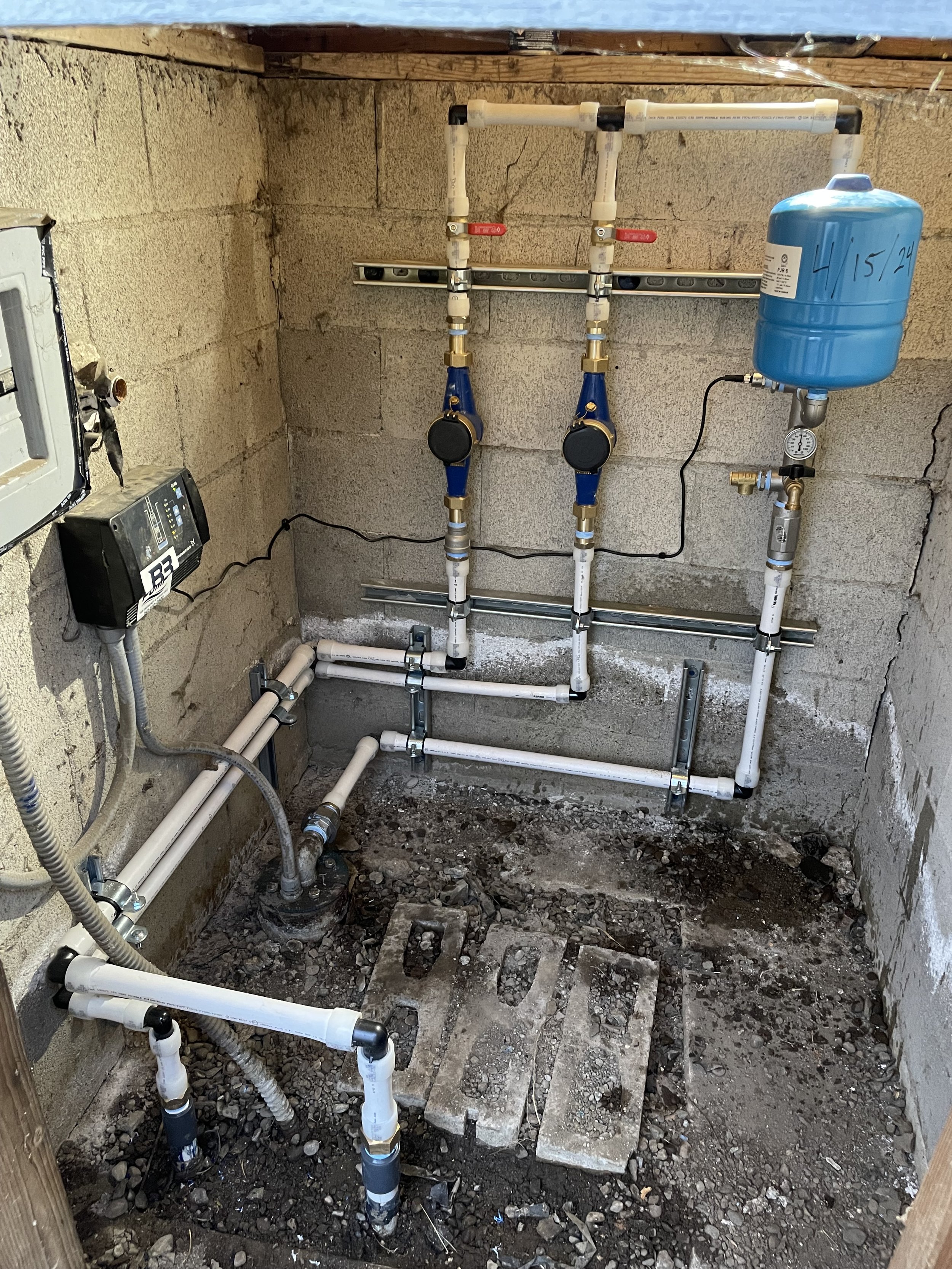 A well plumbing system with white pipes, blue valves, a pressure tank marked with the date '4.15.24', and a water meter, installed in a concrete-block wall space.