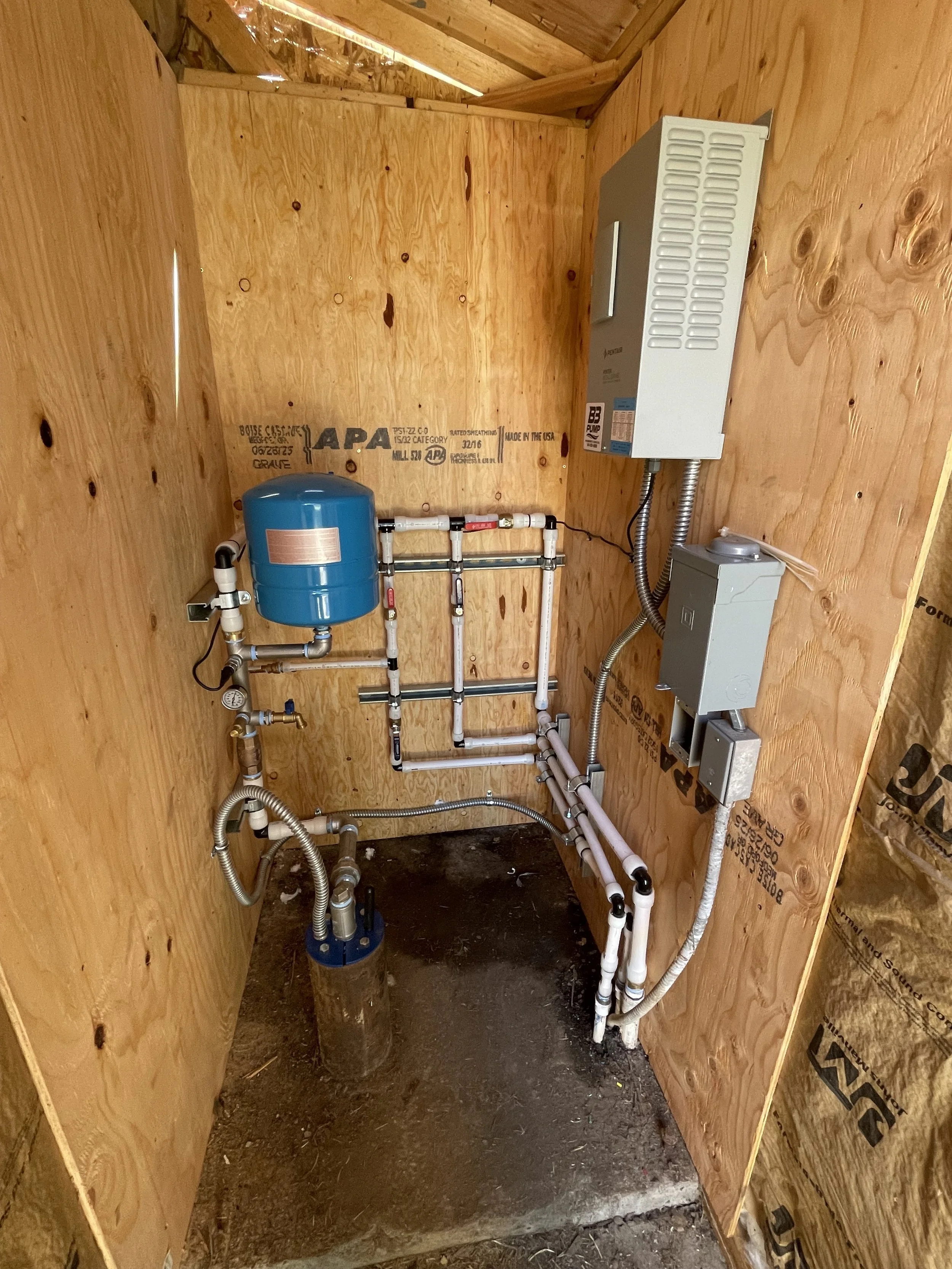 Small pump room with plumbing, a well, VFD, and electrical components, including a blue water pressure tank, various pipes, and electrical boxes mounted on unfinished wooden walls.