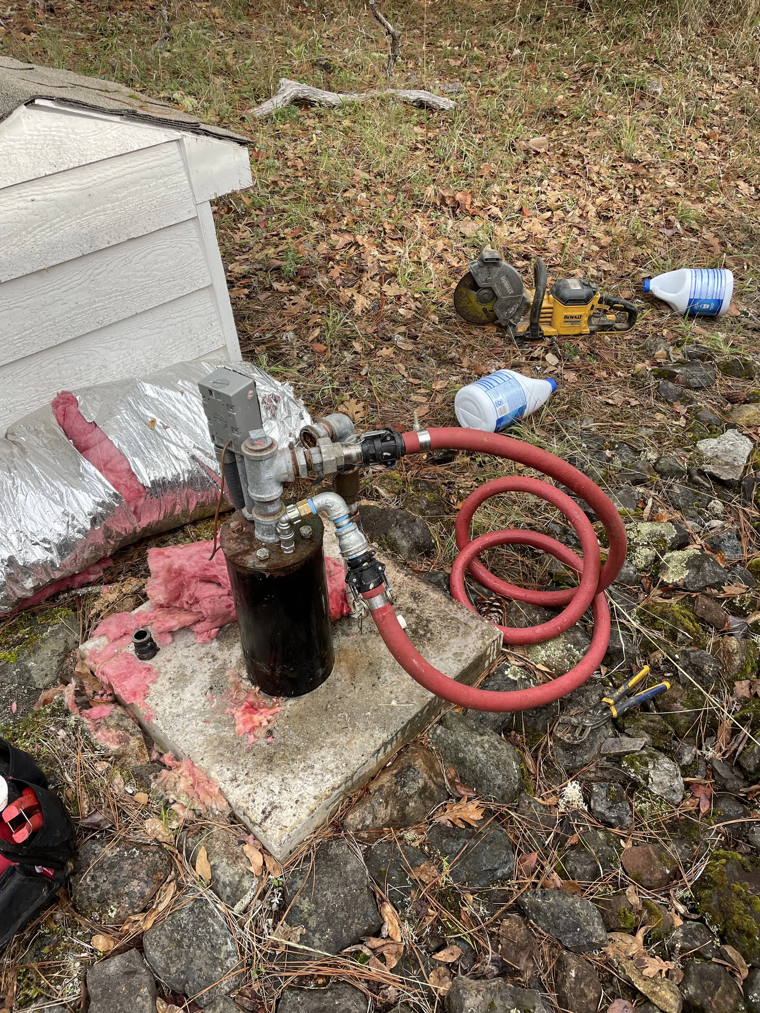 Water well being chlorinated and recirculated, red hose, and tools, situated on a concrete slab near a small white shed with insulation material and surrounded by rocks and fallen leaves.