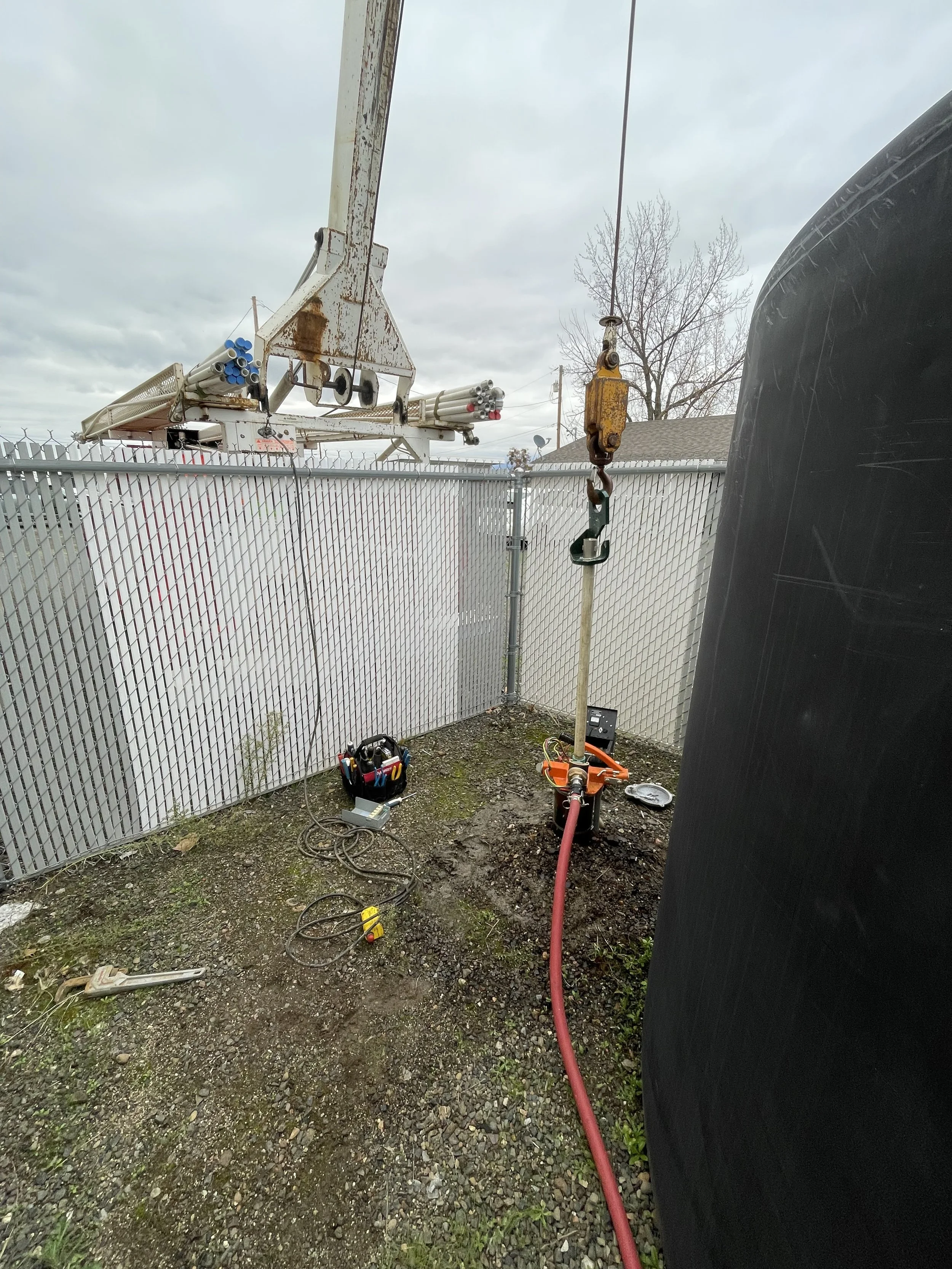 water well pump being pulled with equipment, tools, and a fence, featuring a crane with a hook and a red hose on the ground.