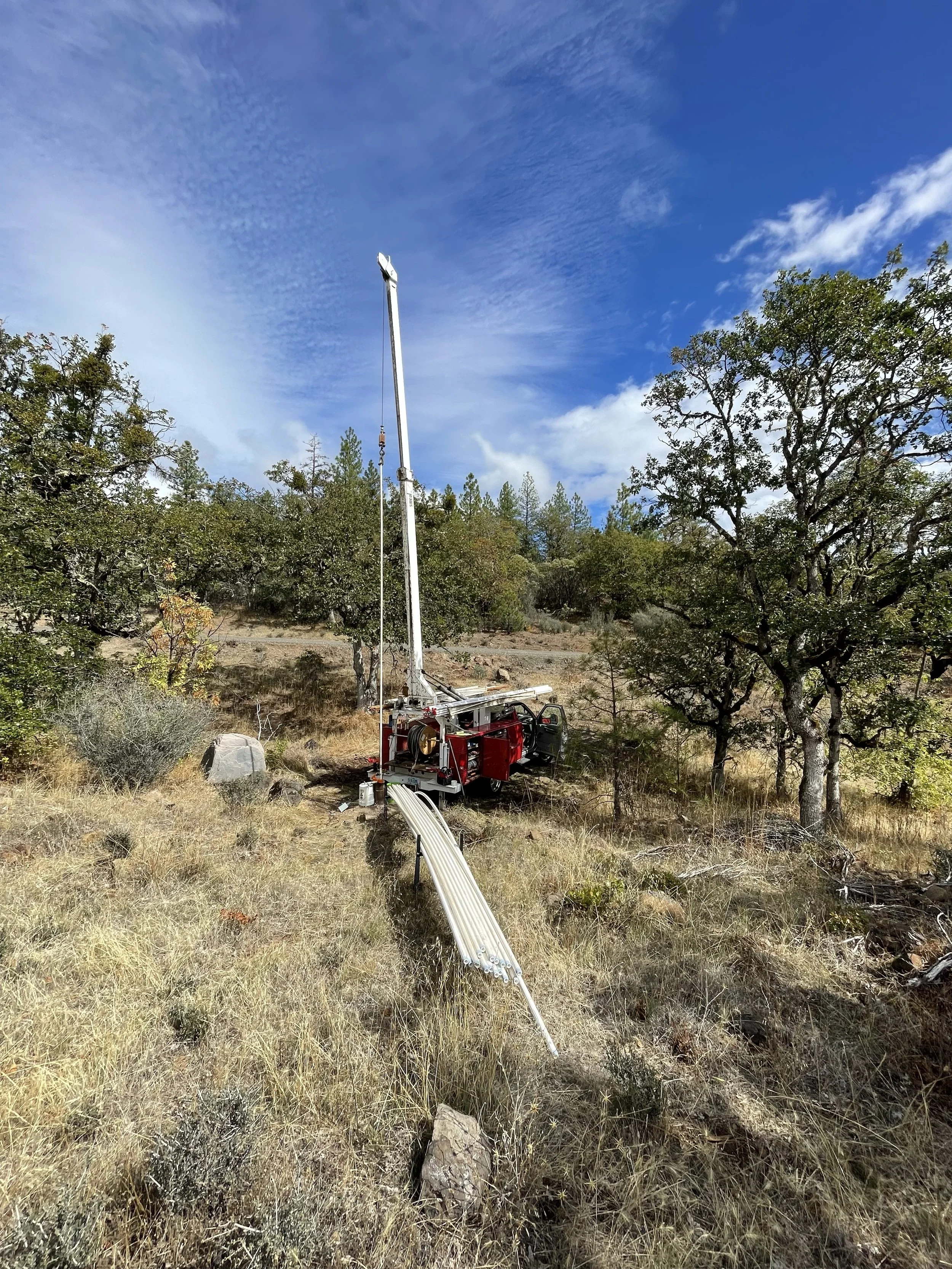 A red well pump hoist truck pulling a well pump on a grassy hillside with trees, rocks, and a partly cloudy sky.