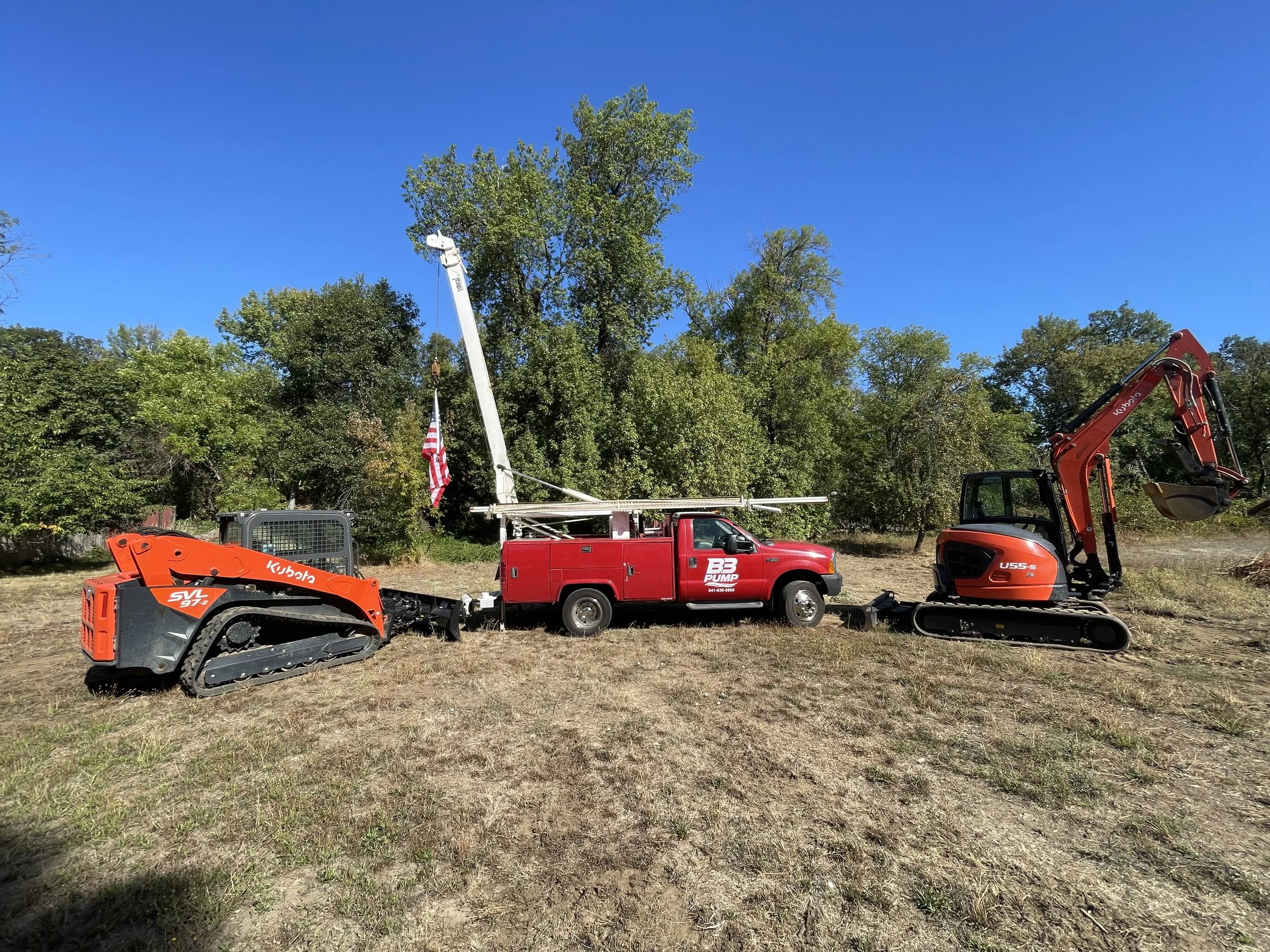 A red service truck with a crane and American flag parked on dry grass, flanked by two small construction excavators, one orange and one red, with green trees and a clear blue sky in the background.