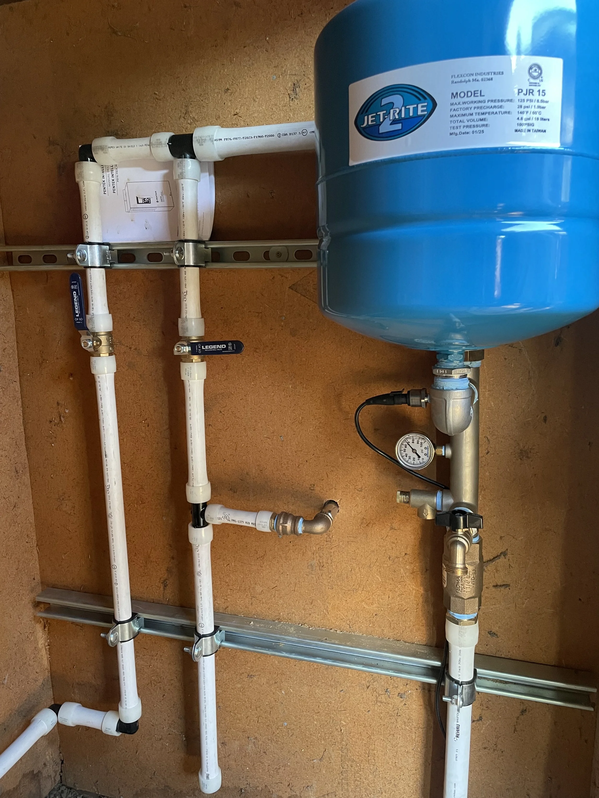 water well Plumbing system with an expansion tank, pipes, valves, a pressure gauge, and a manifold mounted on a brown wall.