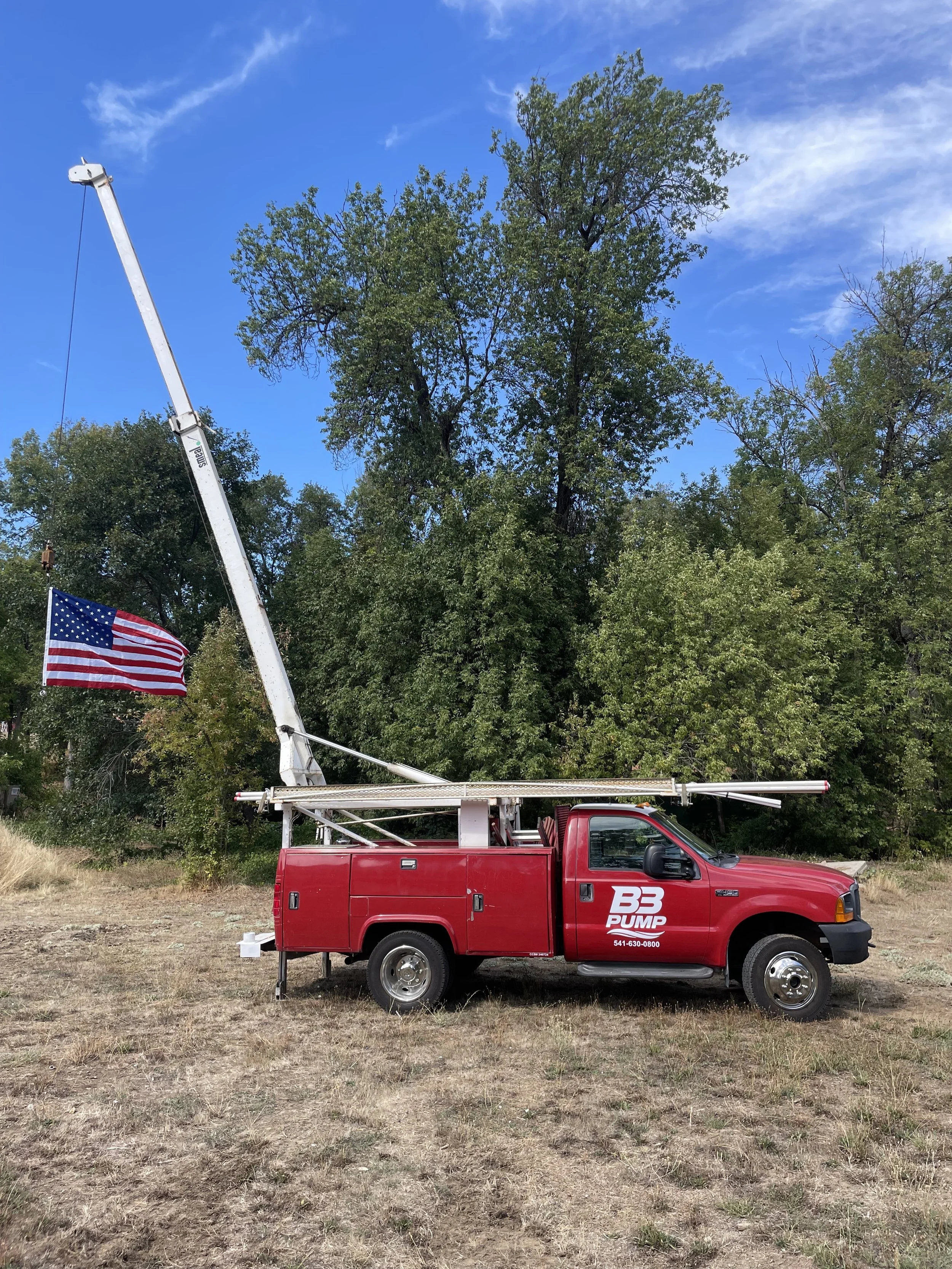 A red well pump hoist truck with an extendable crane and American flag attached, parked on dry grass near trees and blue sky.