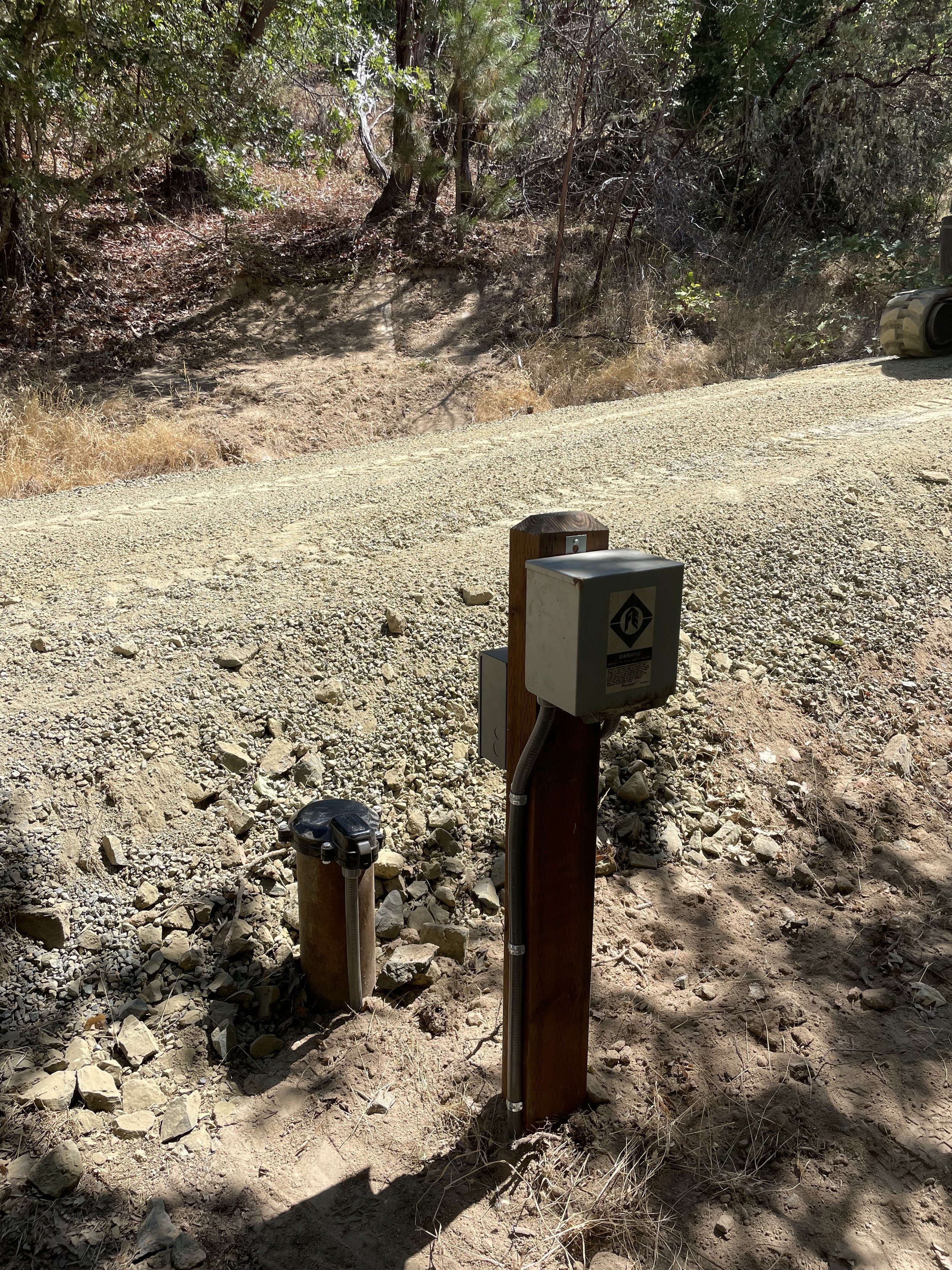 a well head with a pitless cap and a well pump controll box on a wooden post, a small black device on a post nearby, and a car partially visible on the right side.