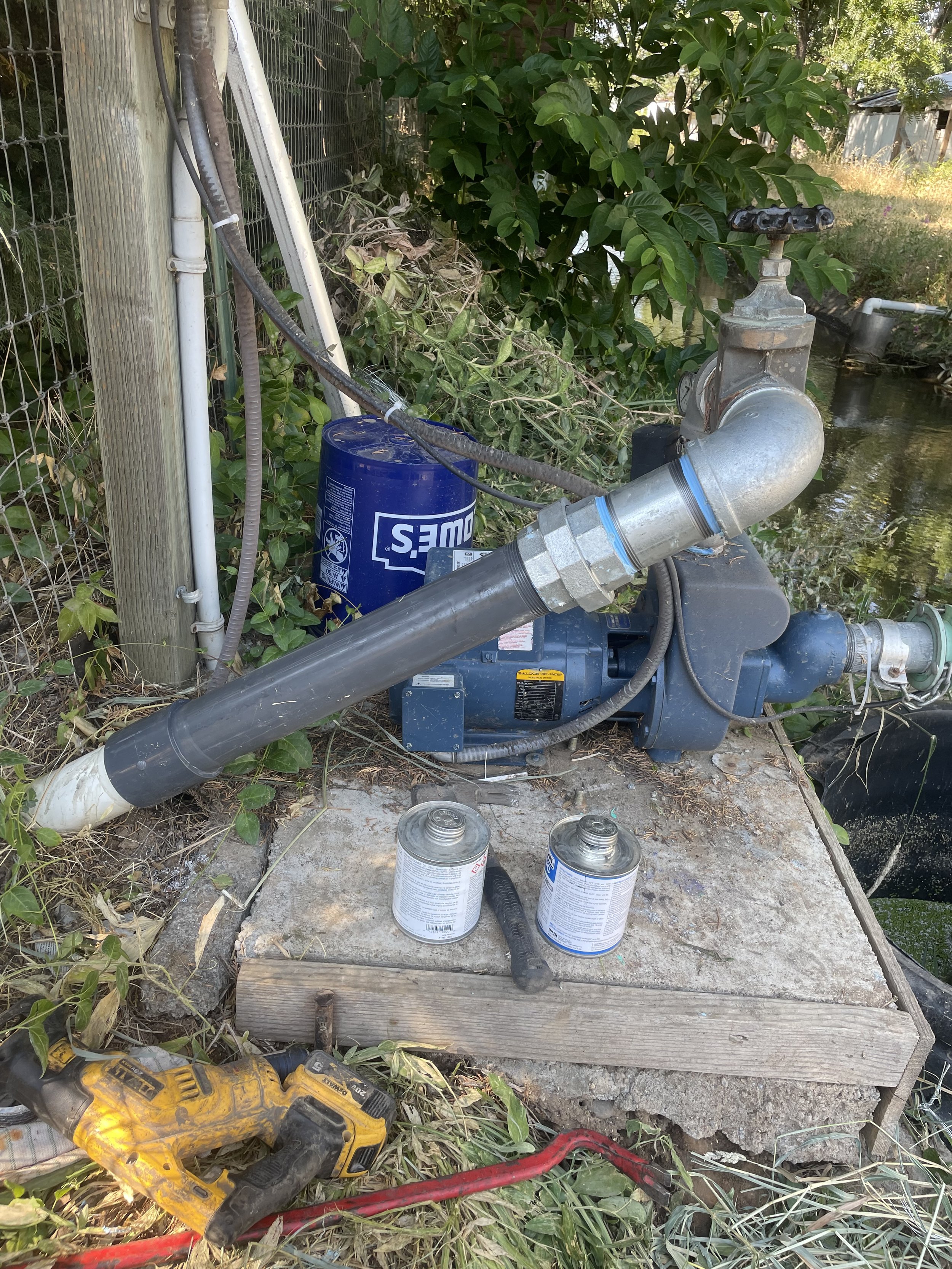 Outdoor irrigation pump system with piping, surrounded by vegetation and tools, on a concrete platform.