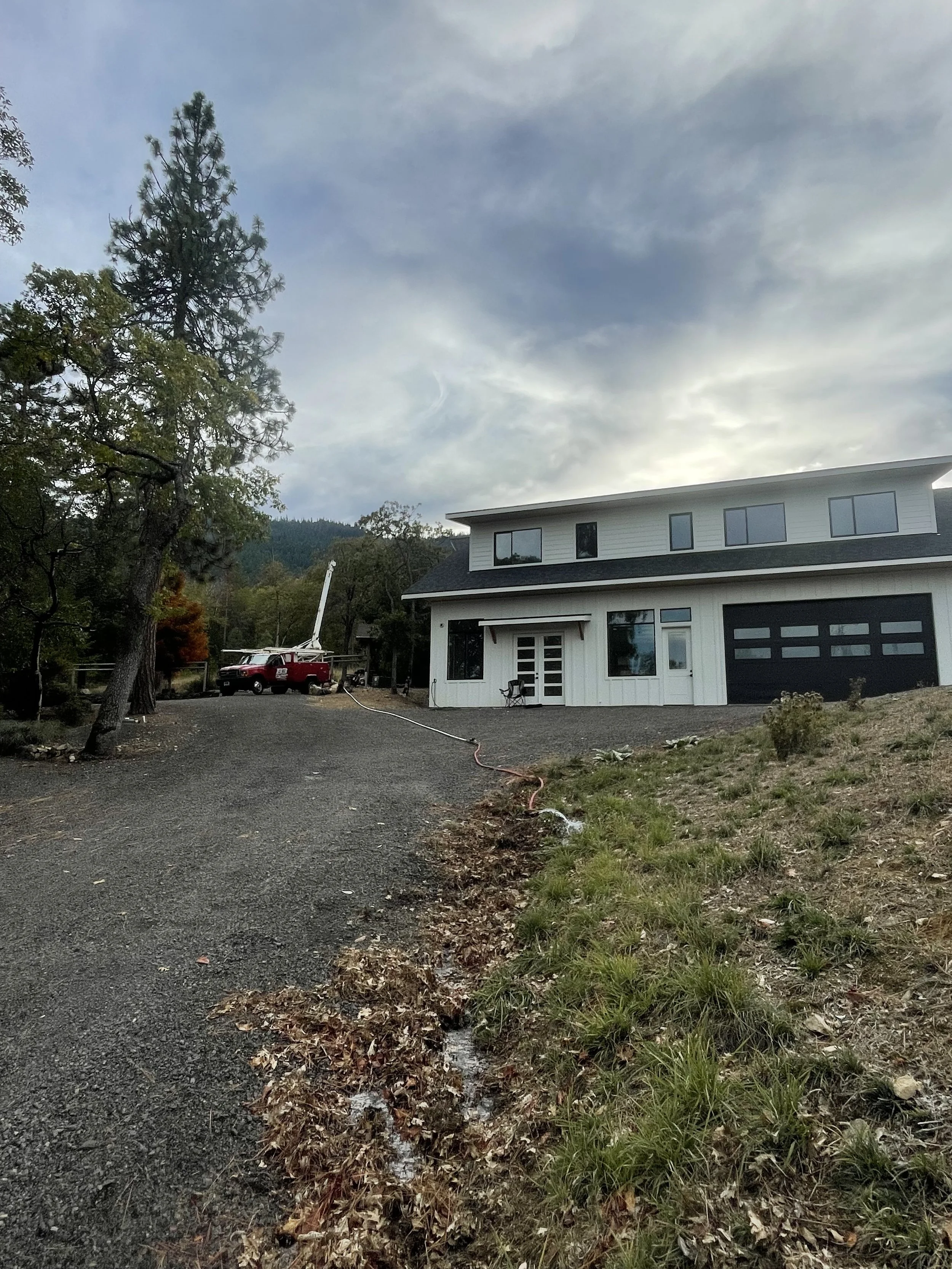 A two-story house with black garage door and large windows, situated on a gravel driveway surrounded by trees and mountains. A red well pump hoist truck with an elevated cherry picker is parked nearby doing a well water flow test